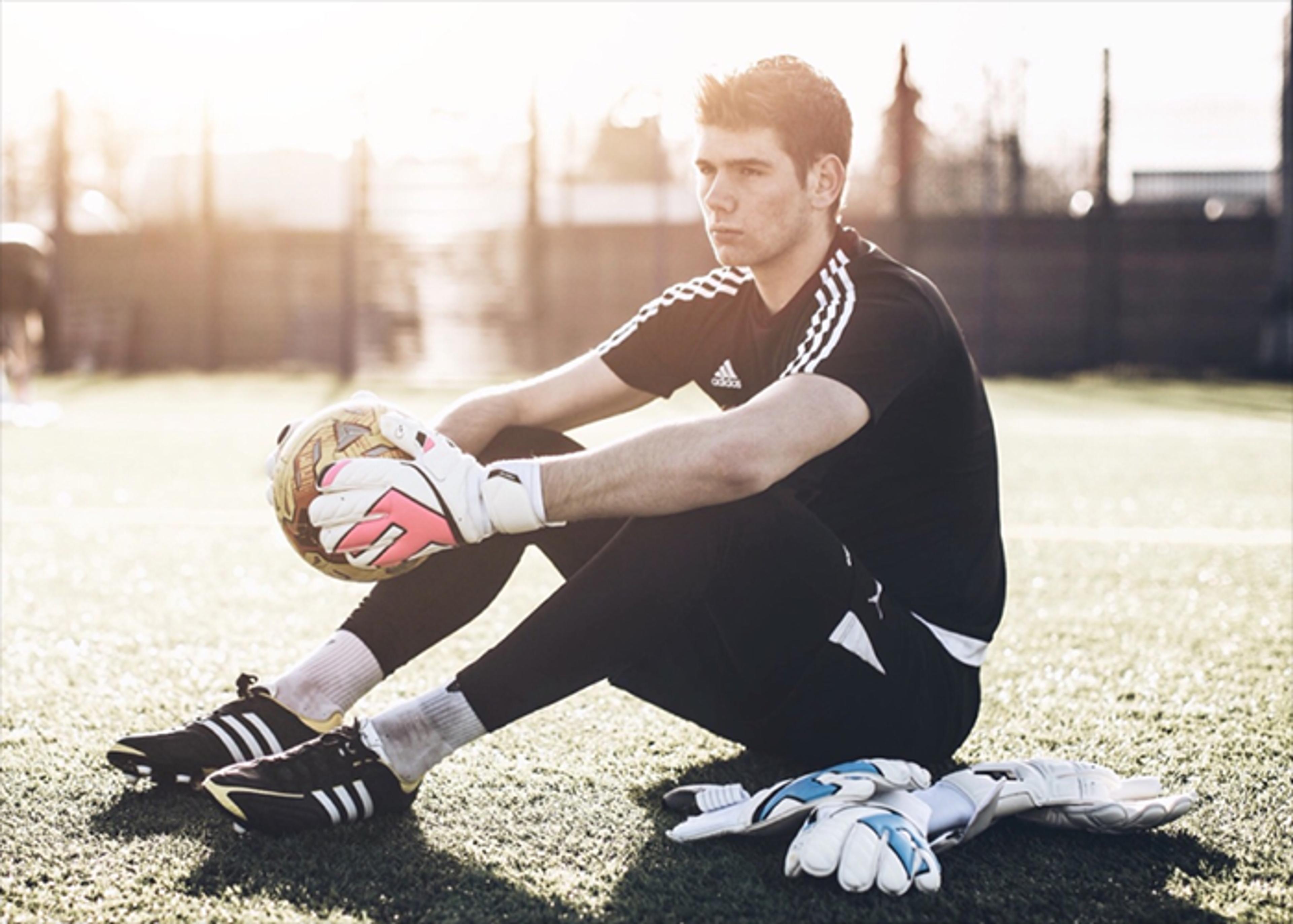 A male soccer goalkeeper in adidas black sportswear and gloves sits on a field holding a ball, with spare gloves on the ground beside him in sunlight.