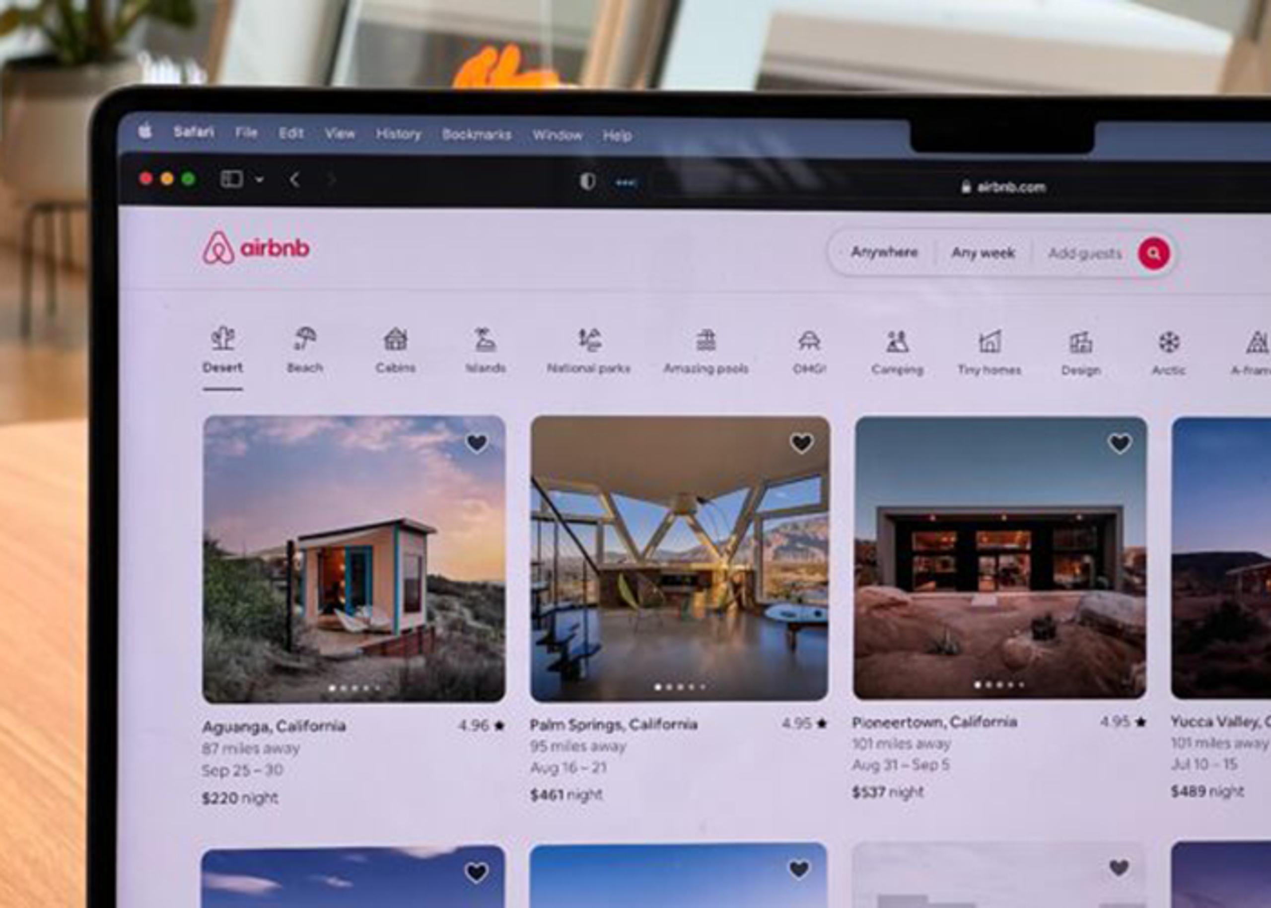 A laptop screen displaying the Airbnb website. It features listings for vacation rentals in locations like Aguanga and Palm Springs, California, with prices and ratings visible. The background shows a bright, modern interior.