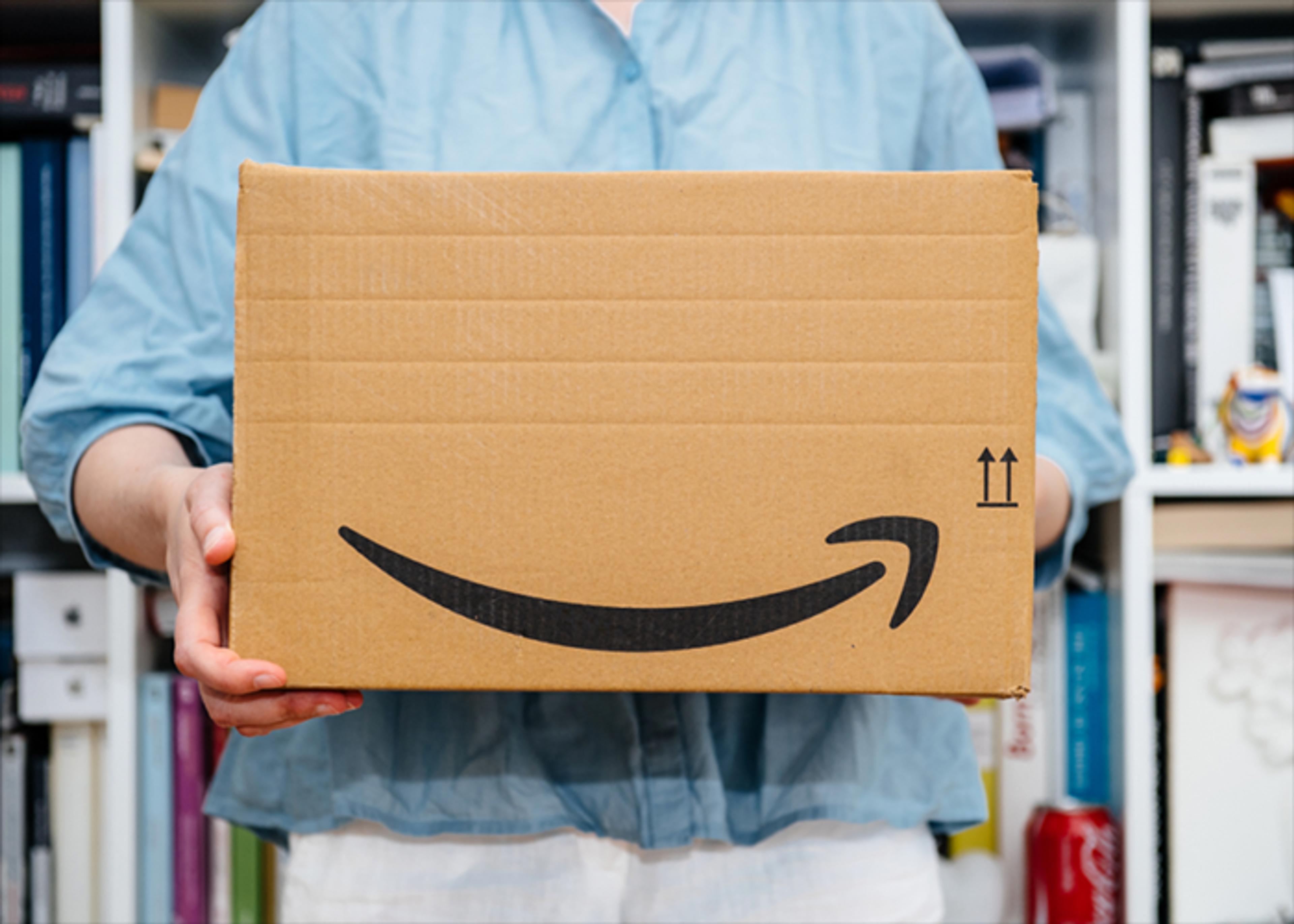A person in a light blue shirt holds a cardboard Amazon package in front of a bookshelf.
