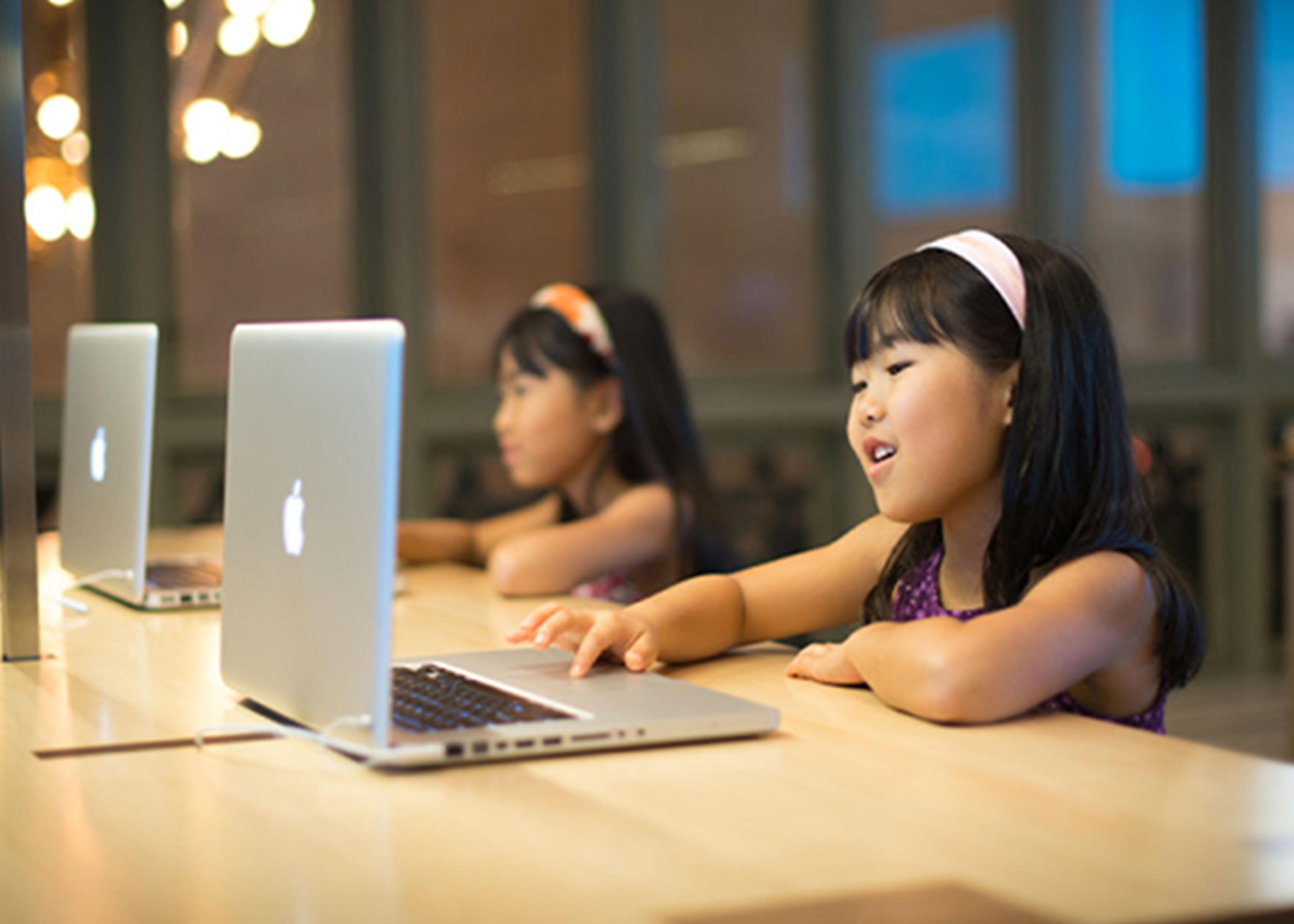 Two girls with long black hair and headbands using laptops at a wooden table in a modern setting. Soft lighting is visible in the background, creating a warm atmosphere.