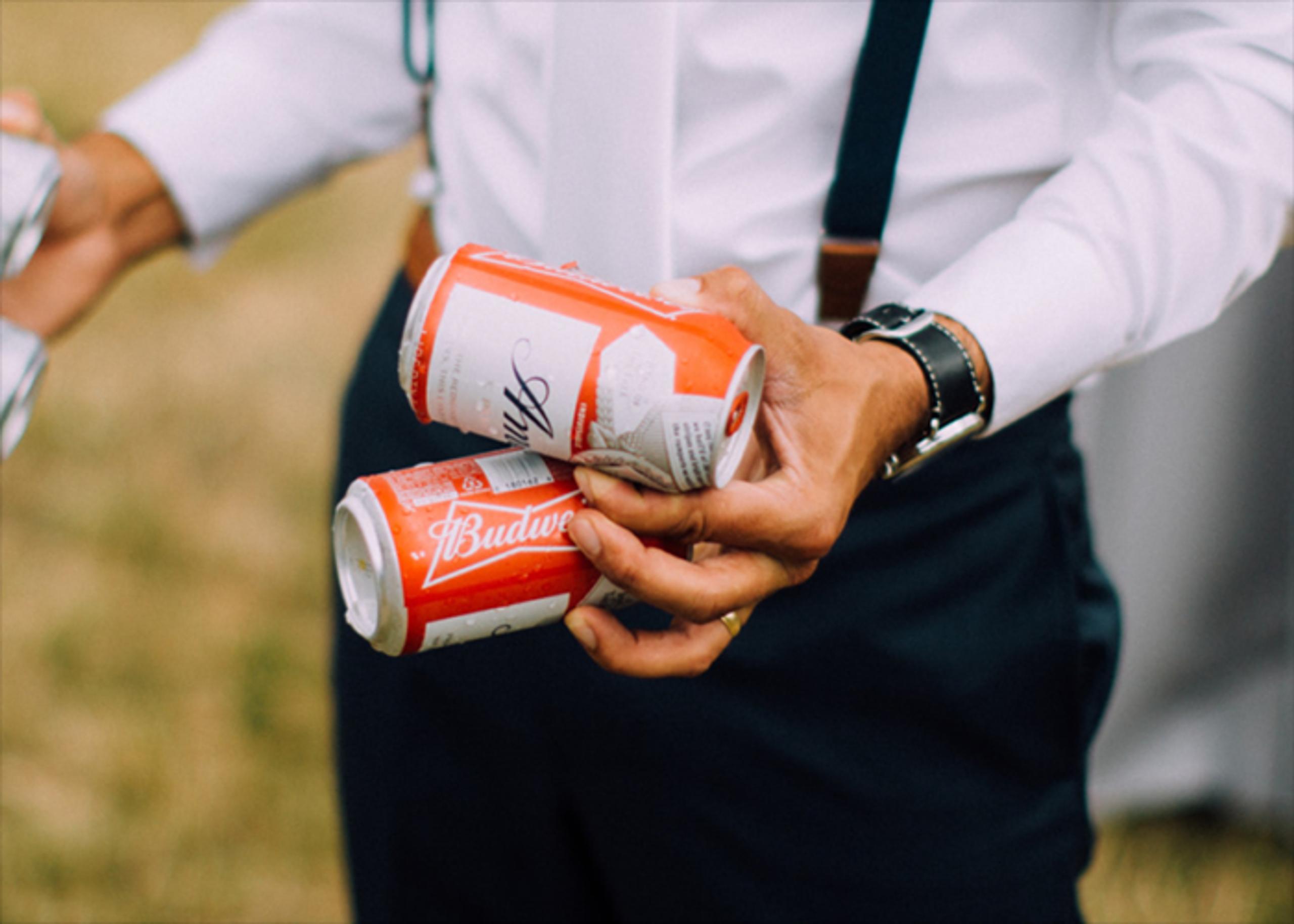 A person holding two red cans of Budweiser in their right hand, wearing a white dress shirt with black suspenders and dark trousers.
