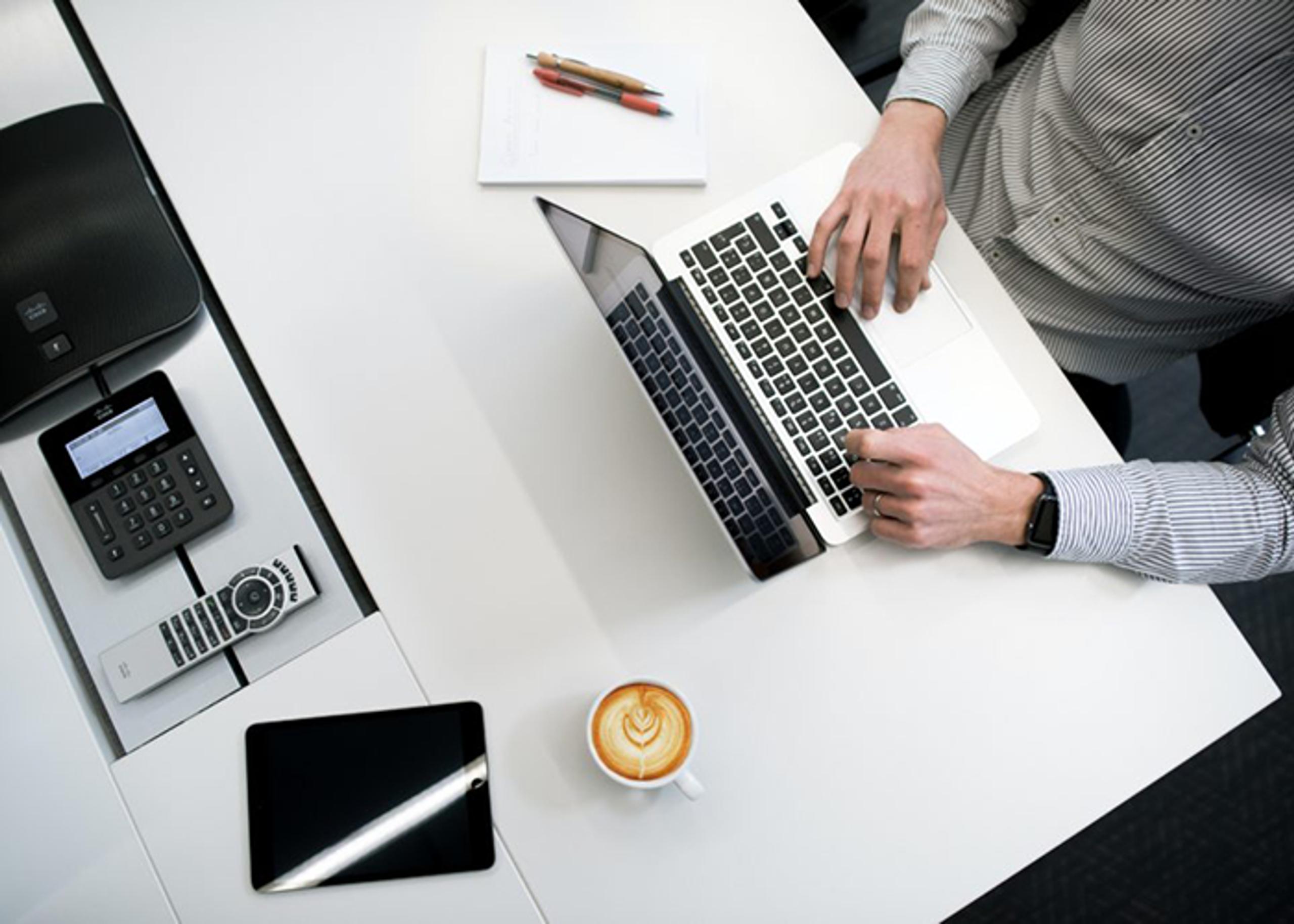 Overhead shot of a man using a laptop at a white table, alongside a tablet, notepaper with pens, a cup of coffee, and conference phone equipment.