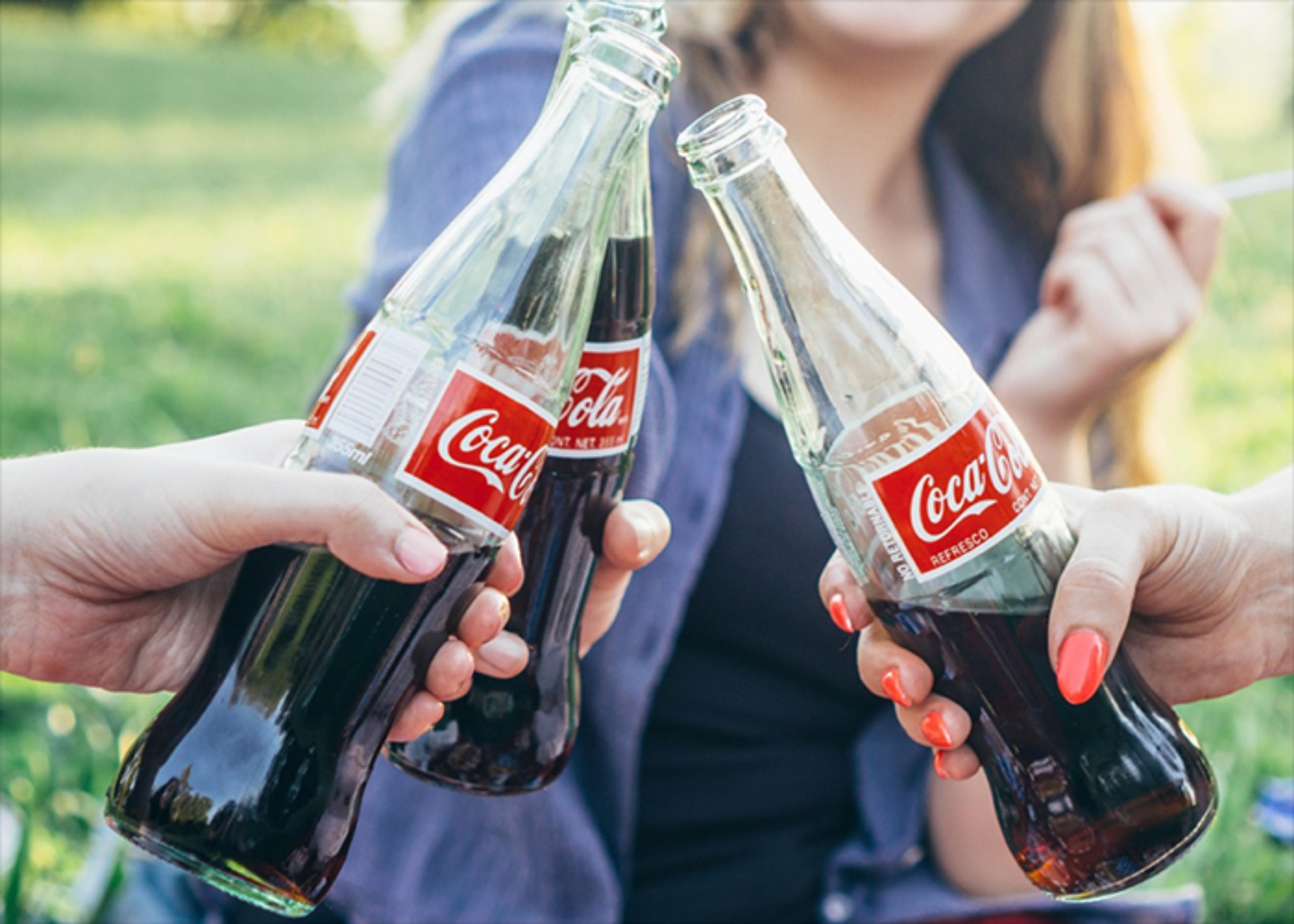 Three hands holding glass bottles of Coca-Cola are raised mid-cheers; a partial view of a woman can be seen in the background.