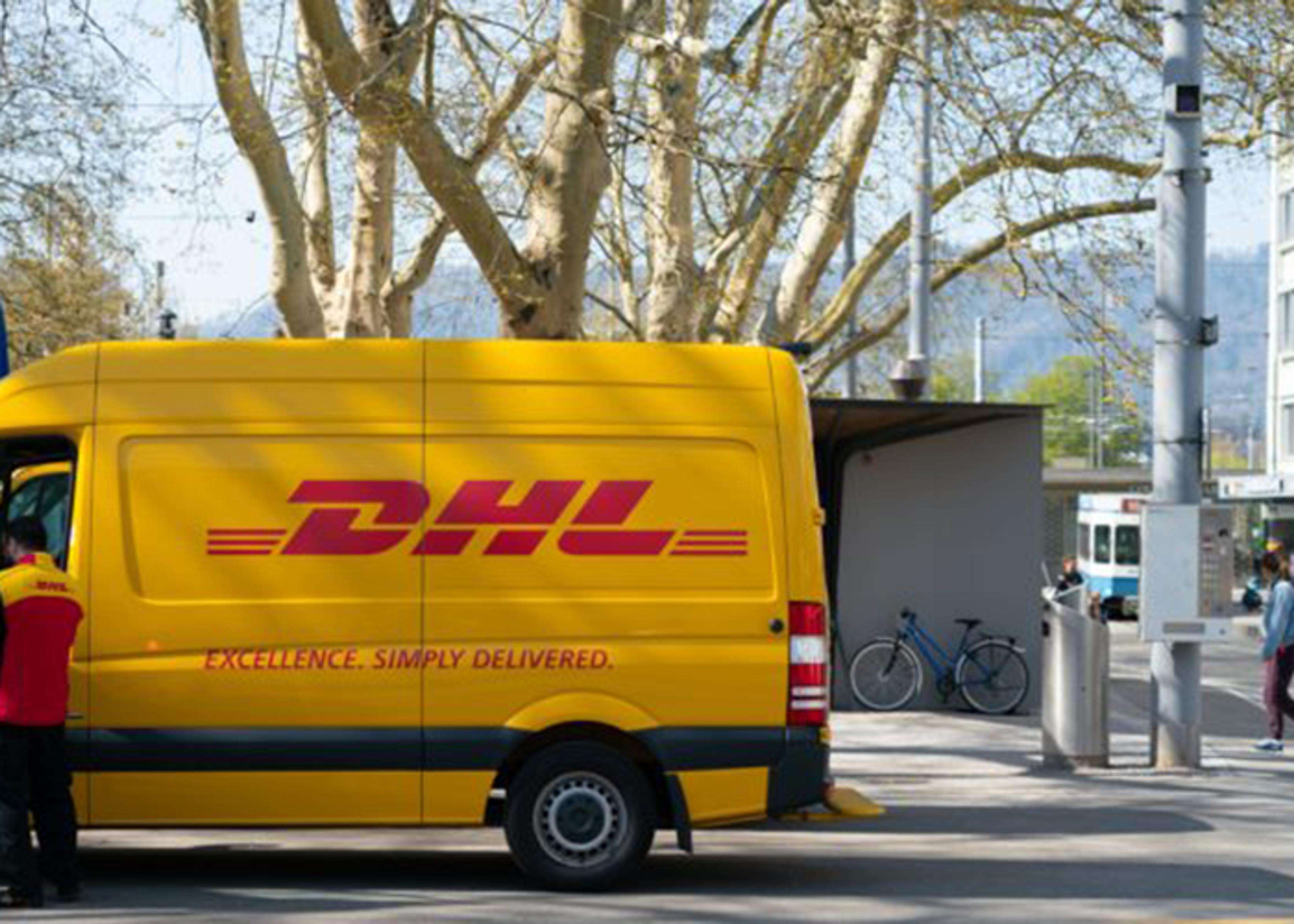 Lateral view of a yellow and red DHL delivery van parked on a city street with bare trees overhead and pedestrians nearby.