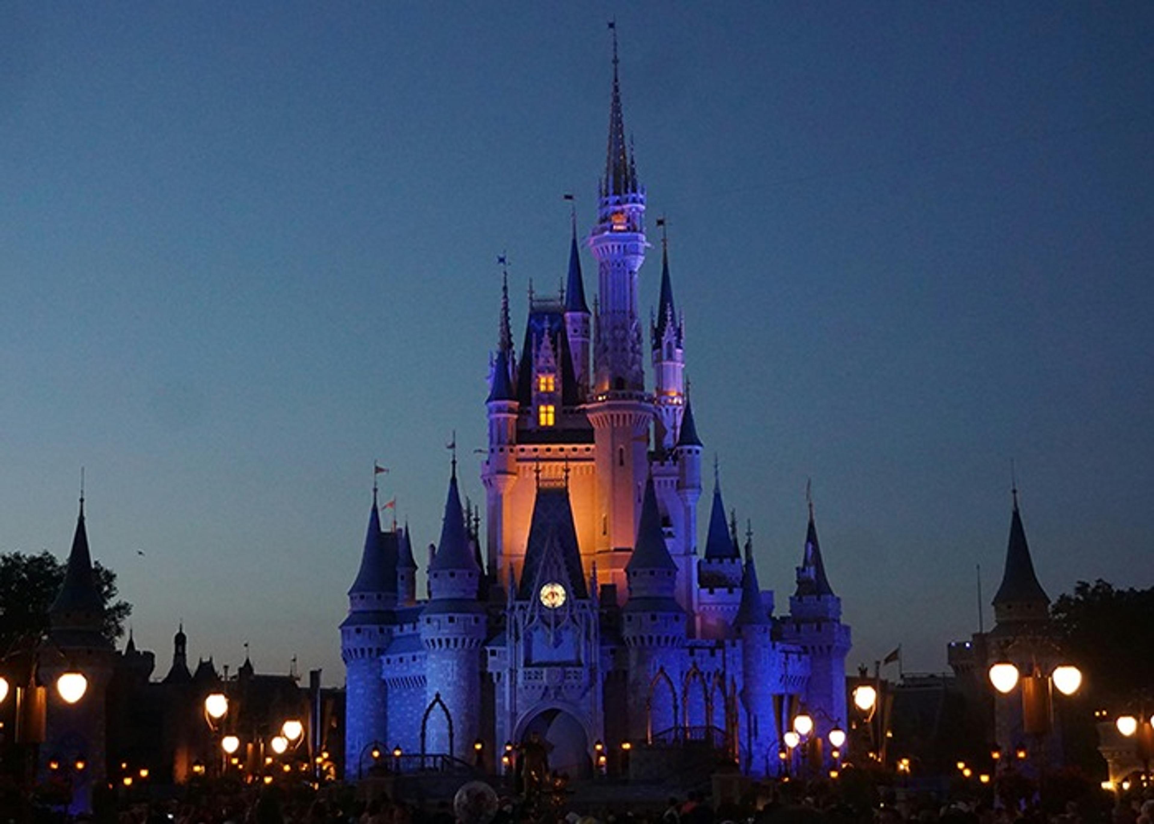 Disney's castle illuminated at dusk, with a crowd of people visible in the foreground.