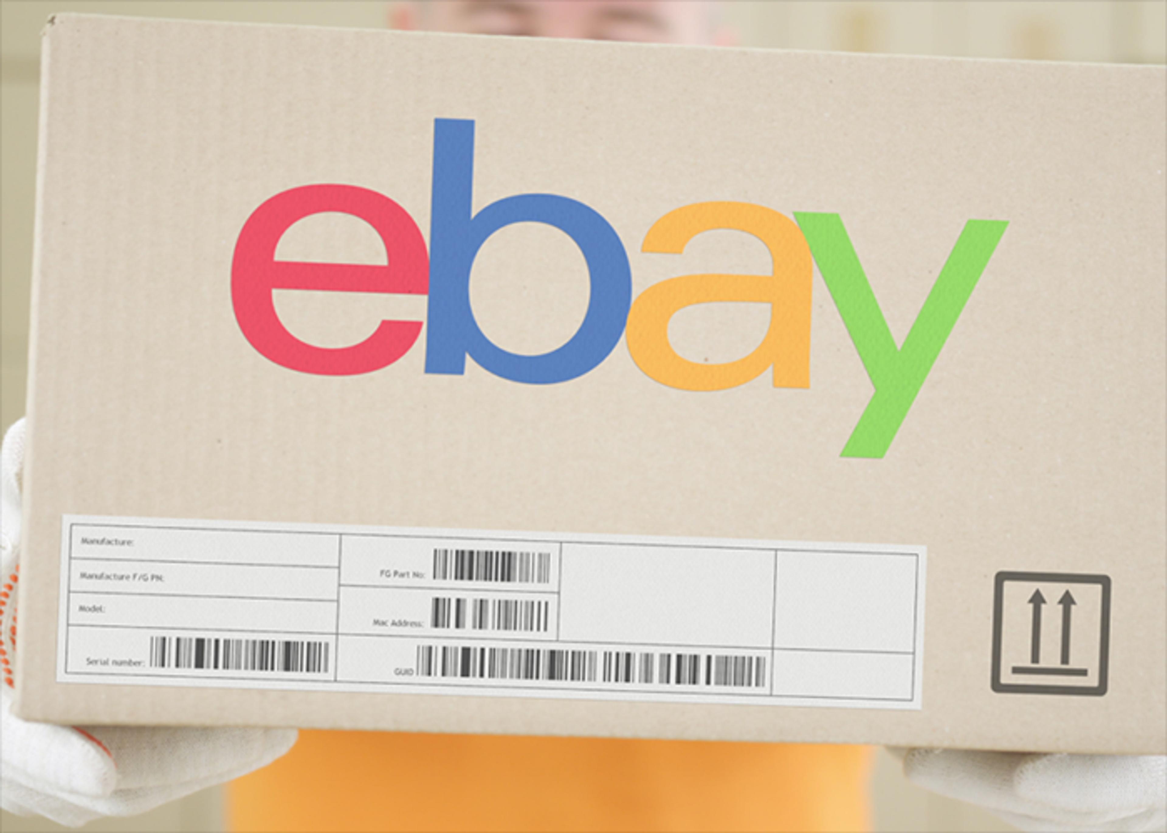 A person wearing an orange shirt and white gloves holds a cardboard box with the eBay logo and shipping labels.