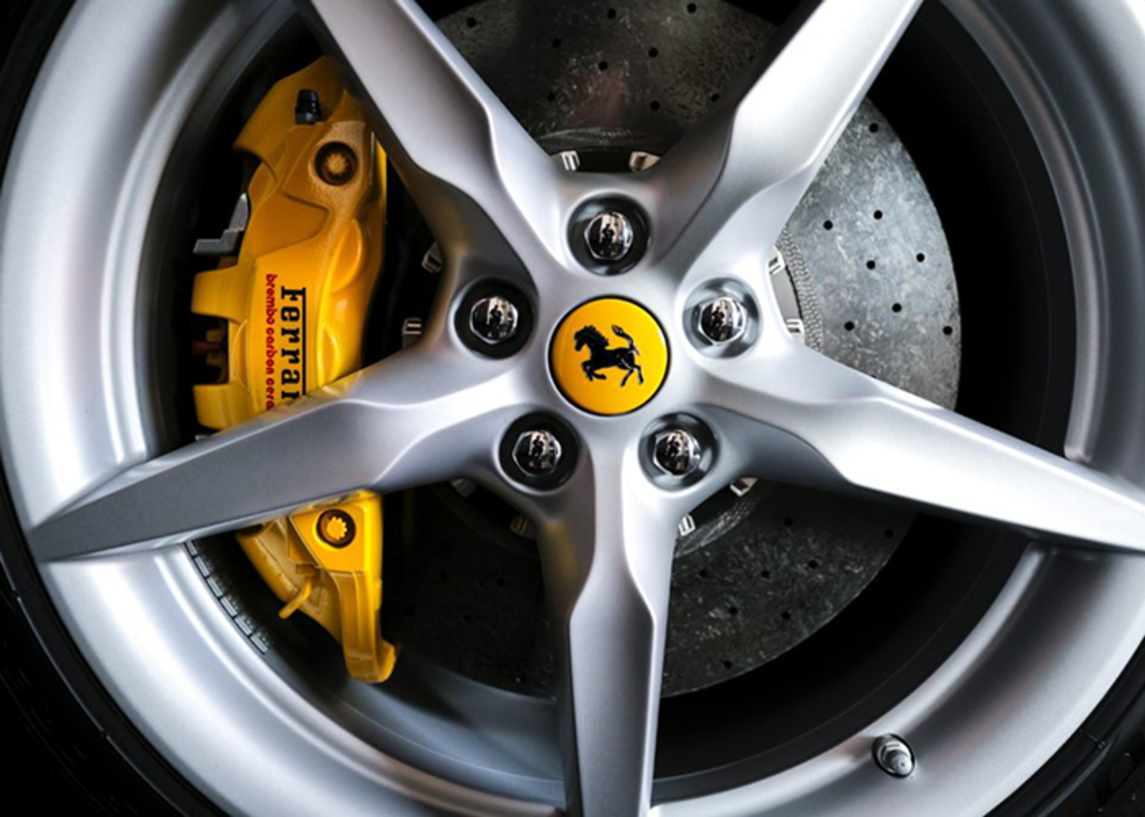 Close-up of a Ferrari wheel showing the silver rim, five black bolts, and yellow prancing horse logo at the center, yellow brake caliper with "Ferrari" and "Brembo carbon ceramic" text, and grey perforated brake disc.