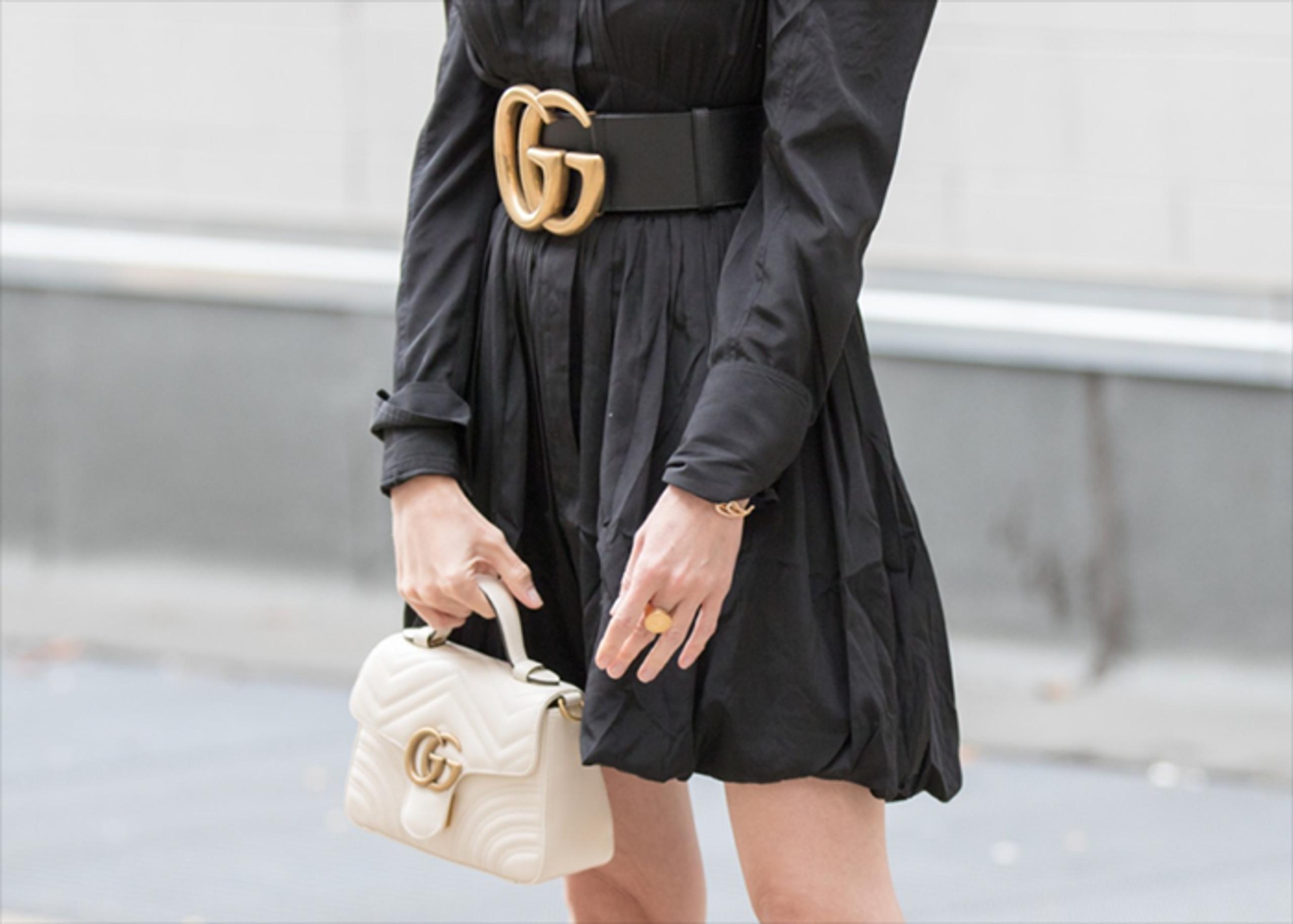 A person wearing a black dress cinched with a large, gold-buckled black belt is holding a small, cream-colored Gucci handbag.