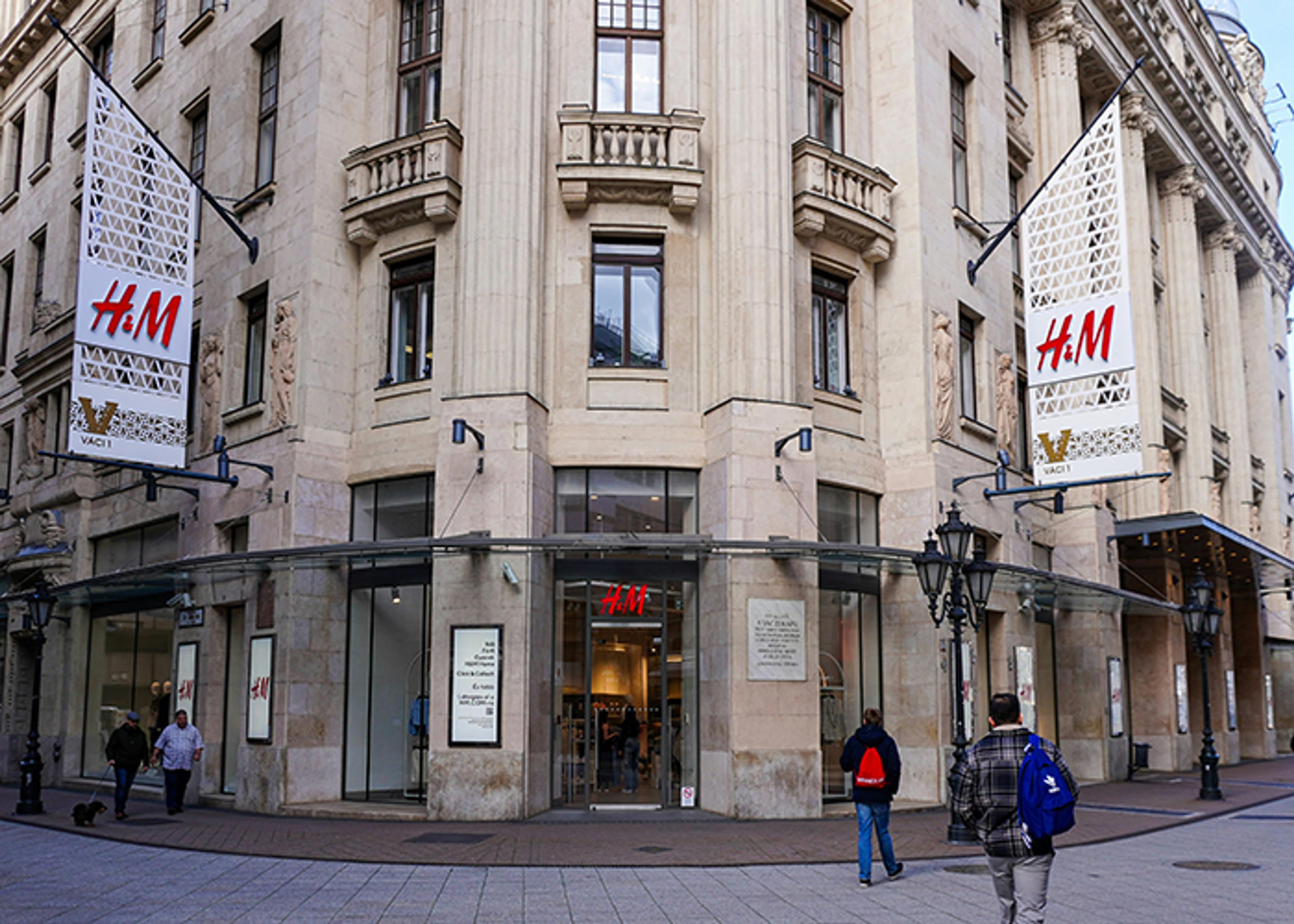 Exterior of the H&M store, featuring beige architectural details and a facade with classical elements; a large H&M logo is visible on a banner, and two pedestrians walk along a sidewalk.