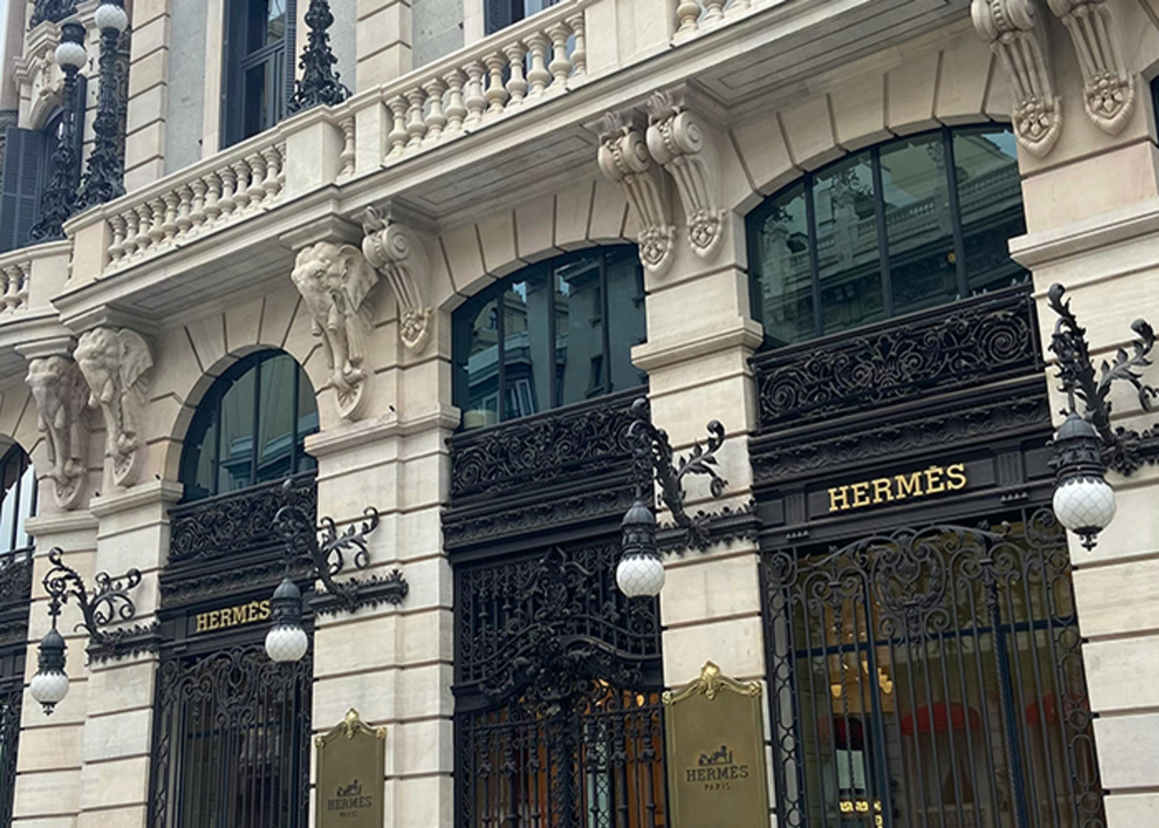 Exterior of the Hermès store, featuring ornate architectural details and signage. The building facade is constructed of light-colored stone, with elephant head supports under an overhanging balcony. Intricate black wrought iron details frame the arched windows and doors. Above each entryway, the word "Hermès" is displayed in gold lettering. Ornate wall mounted lanterns hang on either side of the entrance.