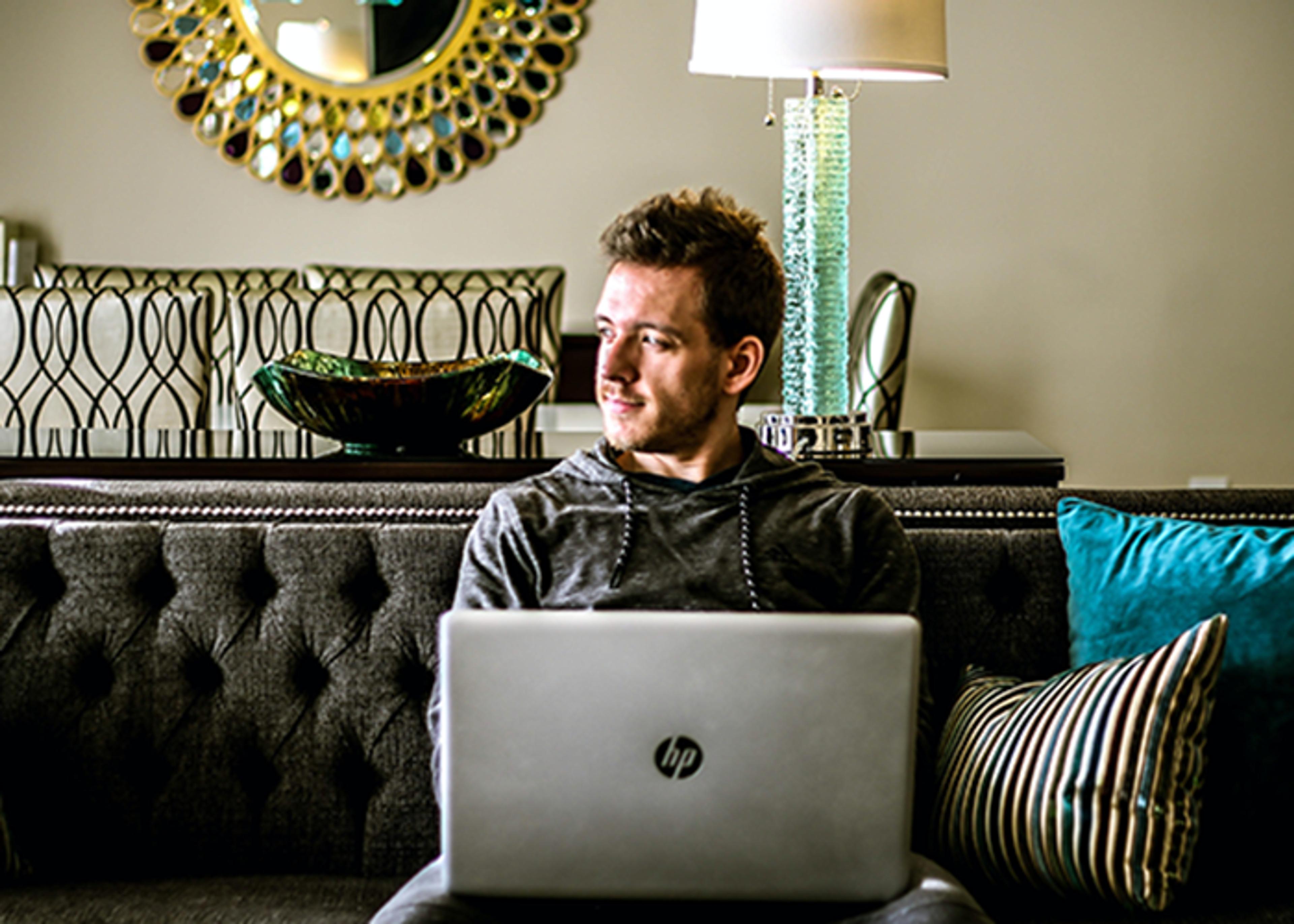 A man sitting on a couch with a HP laptop, looking away with a thoughtful expression.