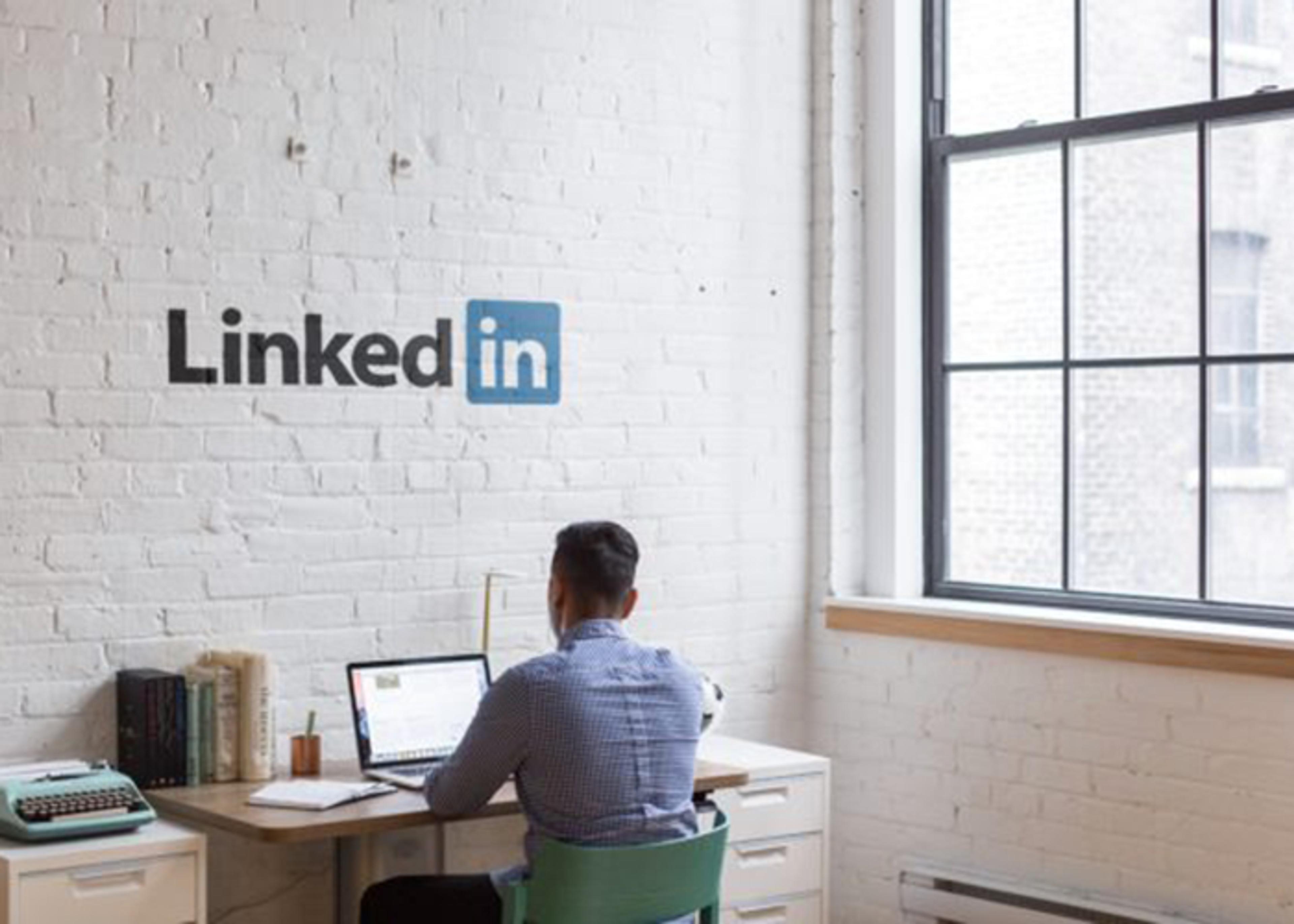 A man sits at a desk, working on a laptop; behind him is a white brick wall with the LinkedIn logo.