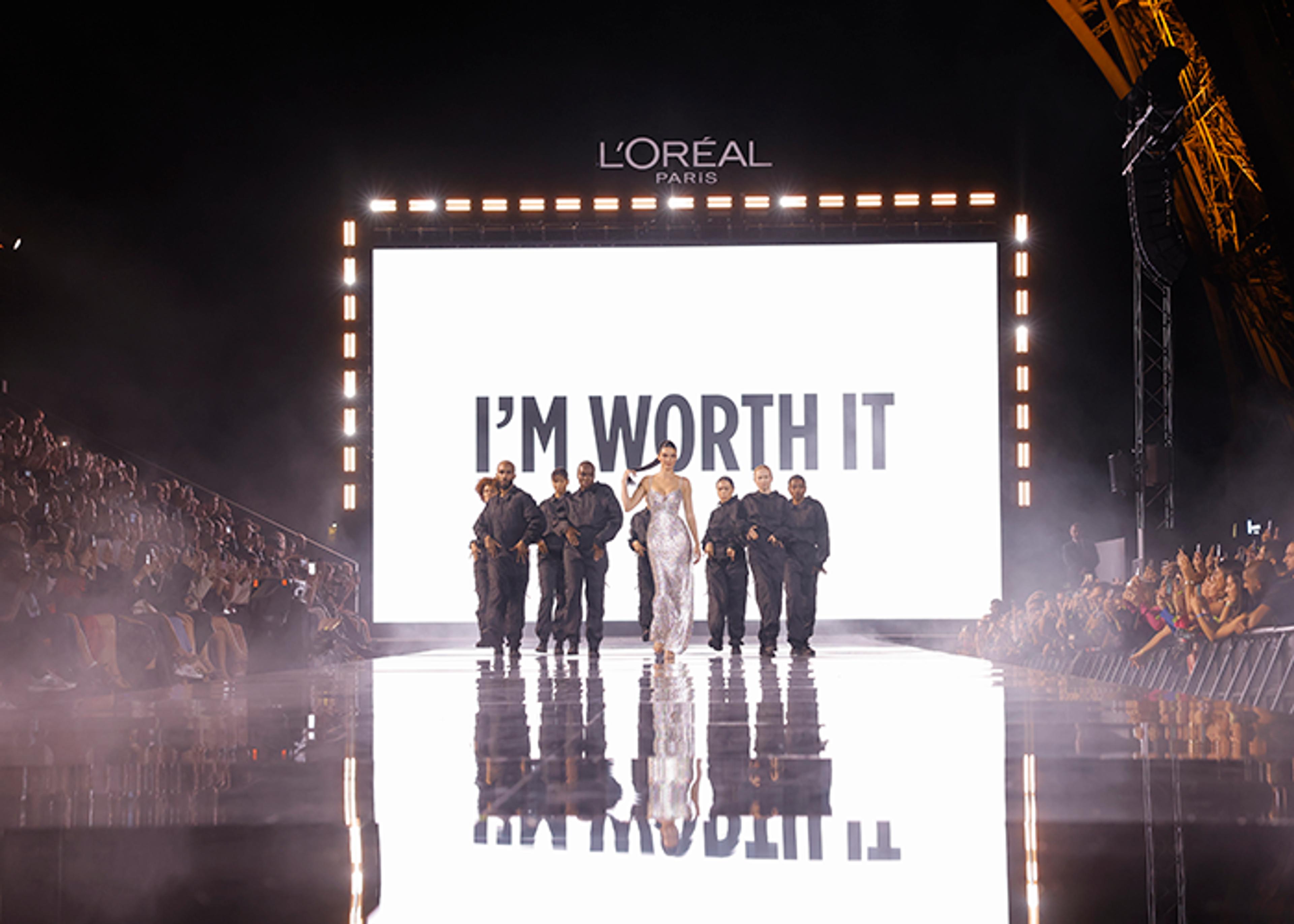 A woman in a sparkling gown walks down a mirrored runway surrounded by security guards beneath a screen displaying the L'Oréal Paris logo and the phrase "I'M WORTH IT," with an audience visible on either side and a partial view of the Eiffel Tower in the background.