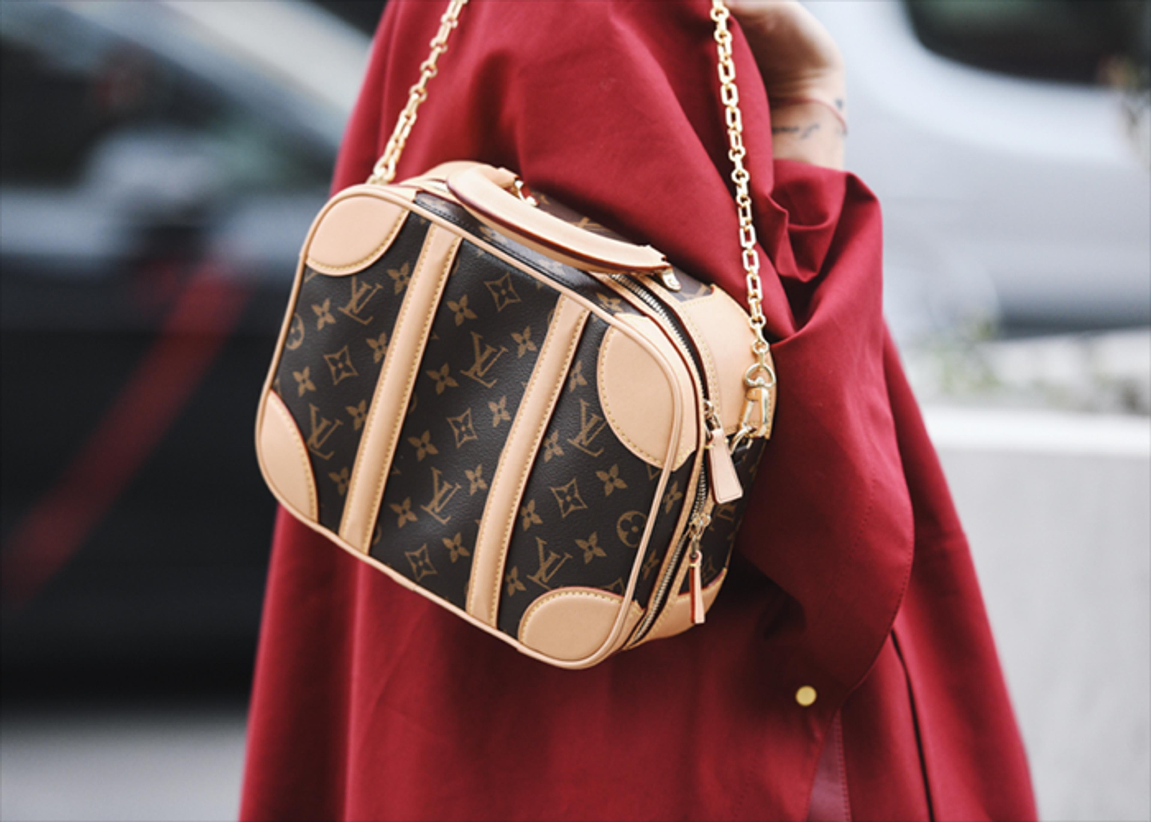 Close-up shot of a brown Louis Vuitton bag being held against a red coat. The bag features a monogram pattern and light tan leather trim, complete with a gold chain strap.