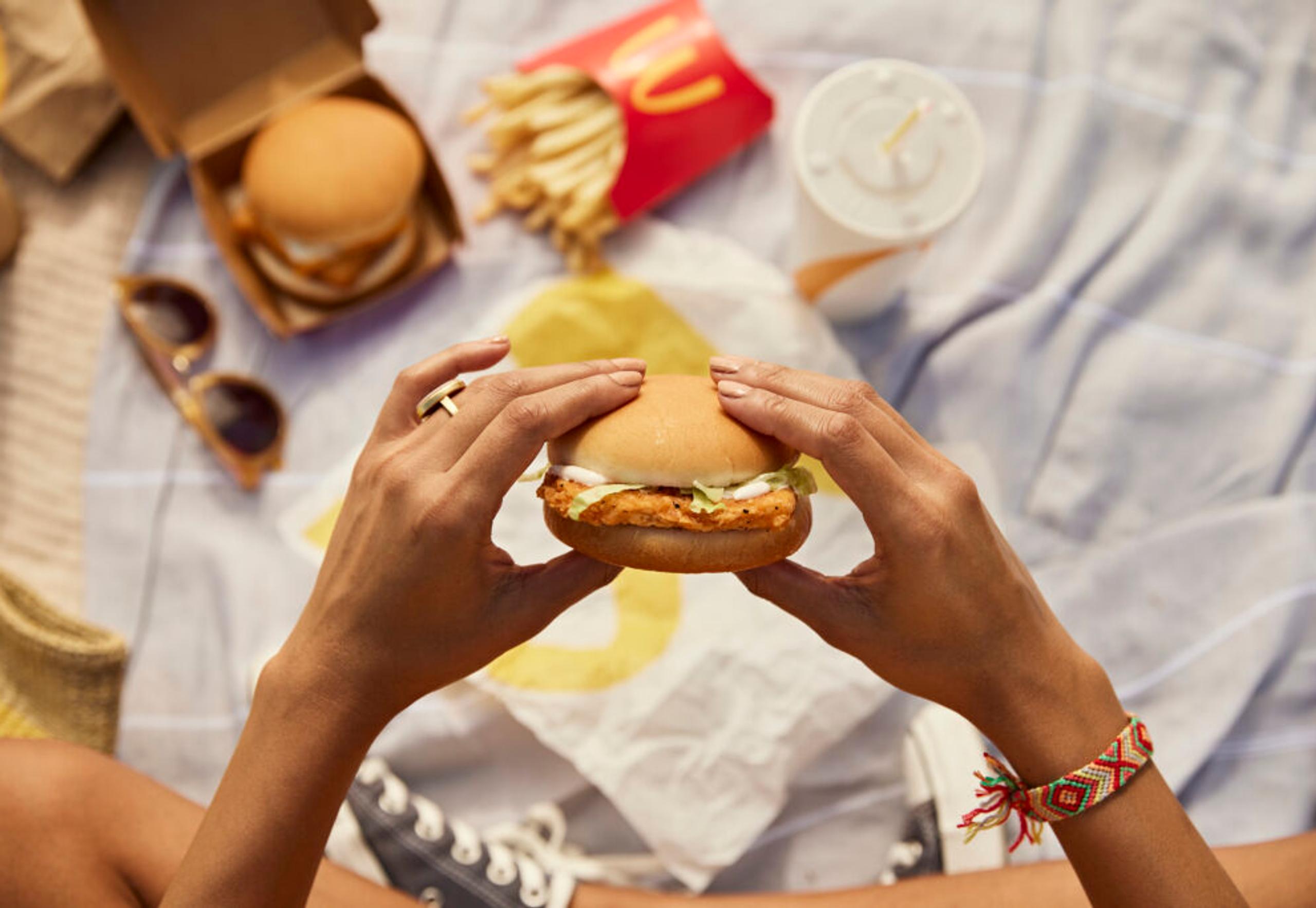 Overhead shot of McDonald's meal on a blanket with a person holding a chicken sandwich topped with lettuce and sauce between their outstretched hands, wearing a ring and a friendship bracelet. The scene contains a red French fry container, a drink with a straw, sunglasses, and a hamburger.