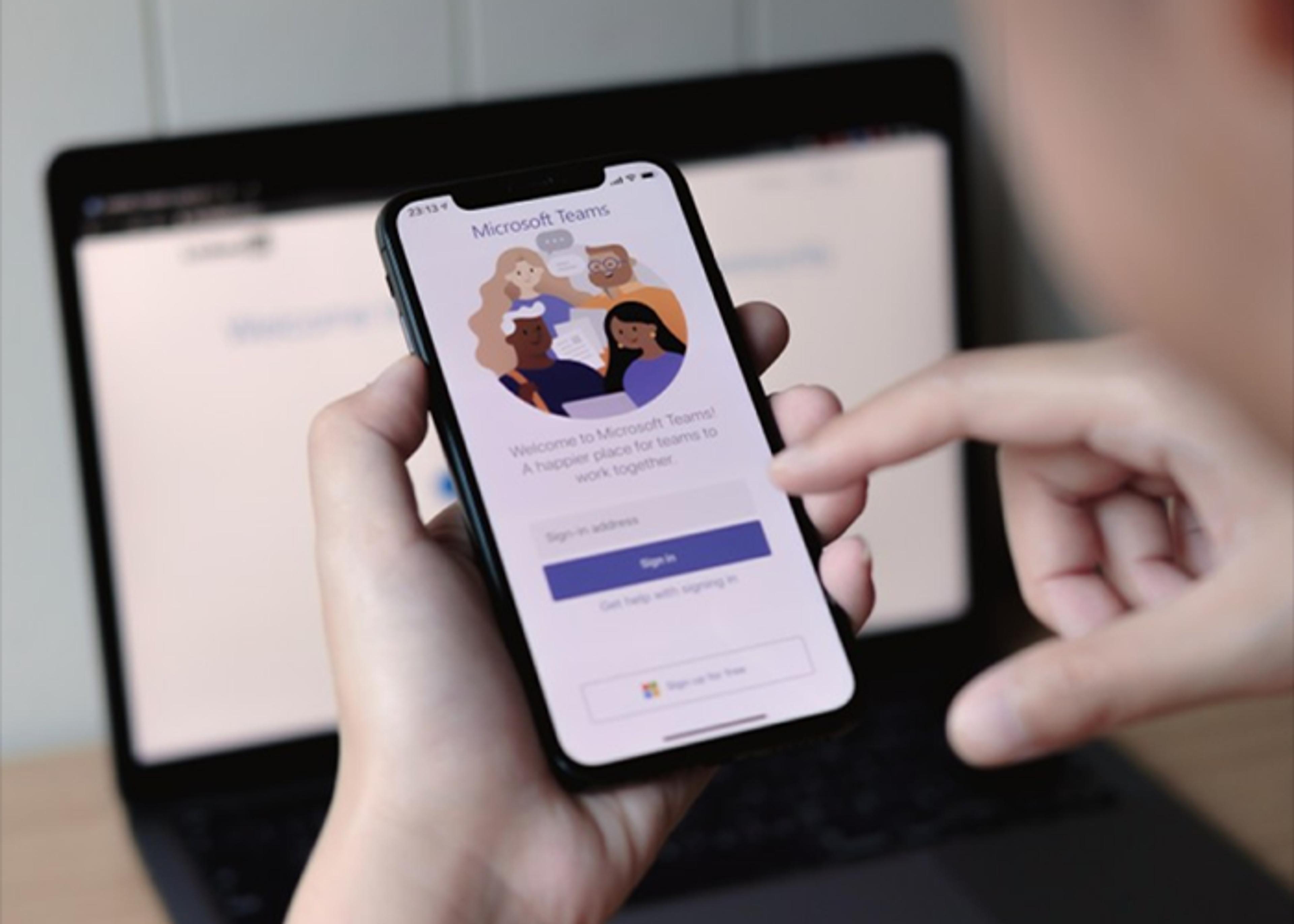 Person’s hand holding a phone displaying the Microsoft Teams login screen, with a blurred laptop in the background.