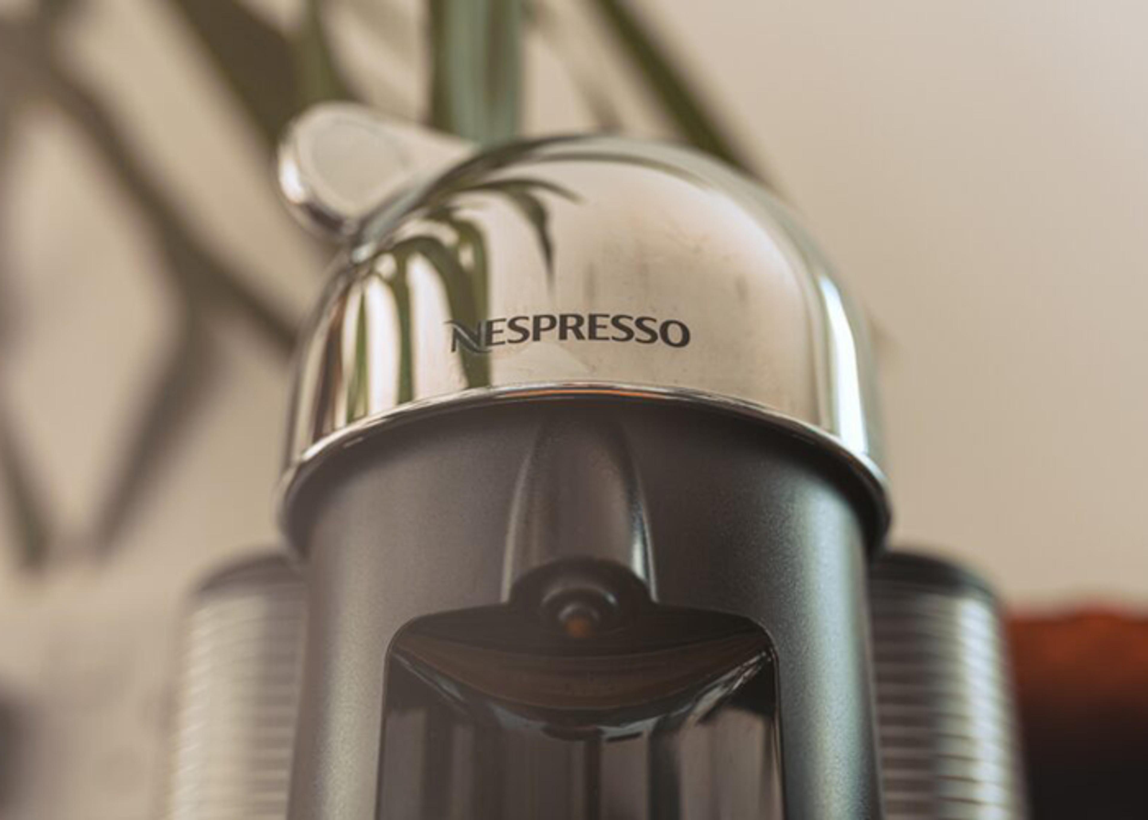 Close-up of a Nespresso coffee machine featuring a polished metal top with the Nespresso logo printed in black, and a gray body, with used capsules partially visible in the background.