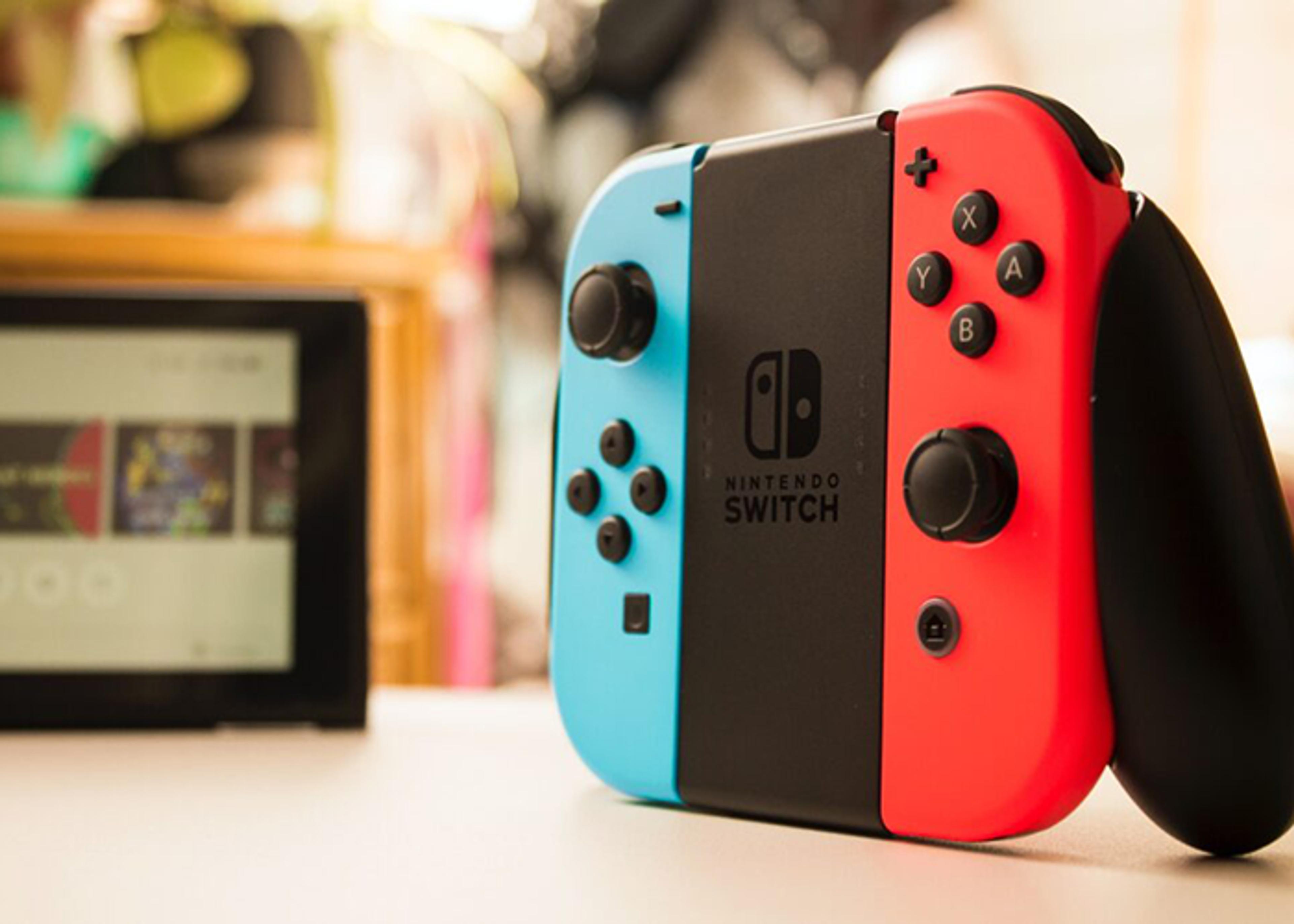 Close-up of a Nintendo Switch console with detached blue and red Joy-Con controllers on a light, neutral surface. The Nintendo Switch screen is visible in the blurred background.