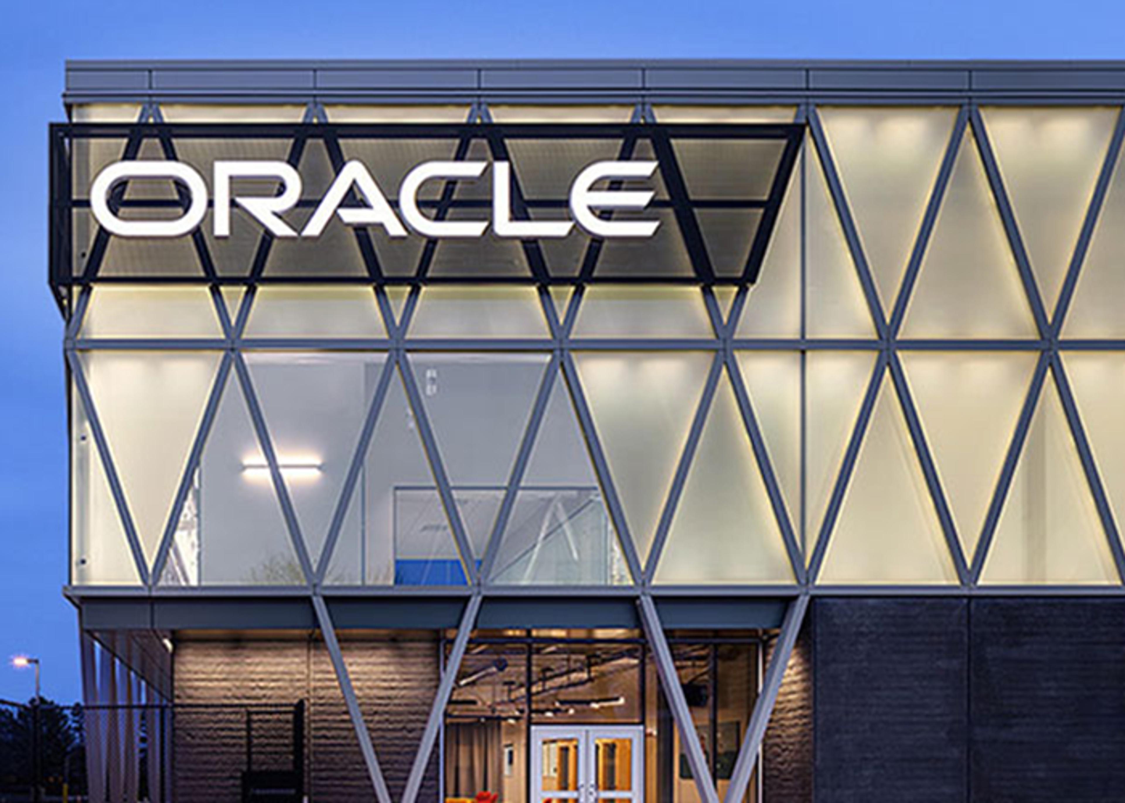 Exterior view of the Oracle building featuring a geometric facade with metal framework forming diamond shapes. The Oracle logo is prominently displayed in illuminated, bold white letters atop the structure. A doorway at the entrance is visible on the ground floor.