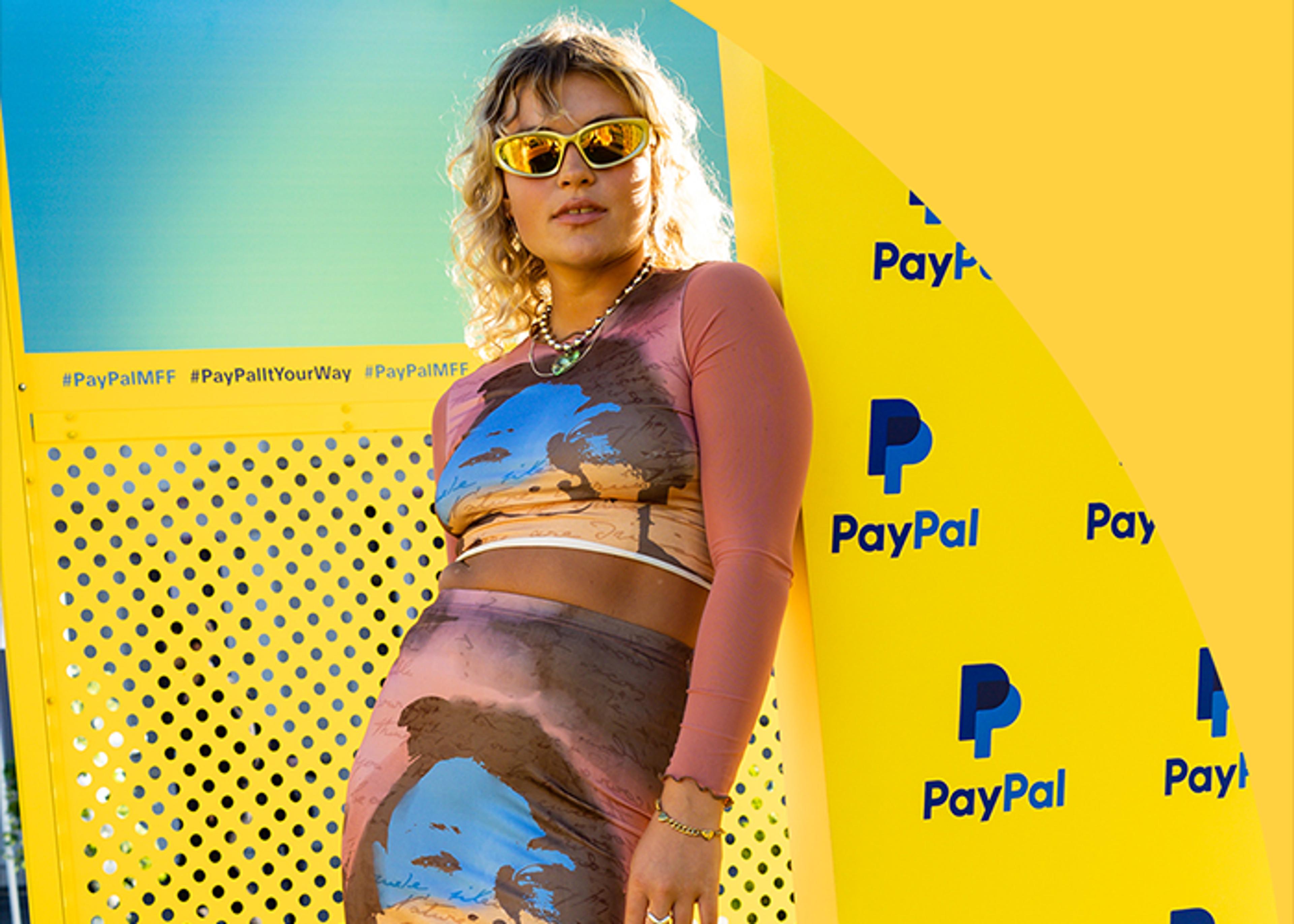 A woman with curly blonde hair and sunglasses leans against a yellow PayPal branded wall with the hashtag #PayPalMFF visible. She wears a long-sleeved top and skirt with a brown and blue scenic print. Her expression is neutral.