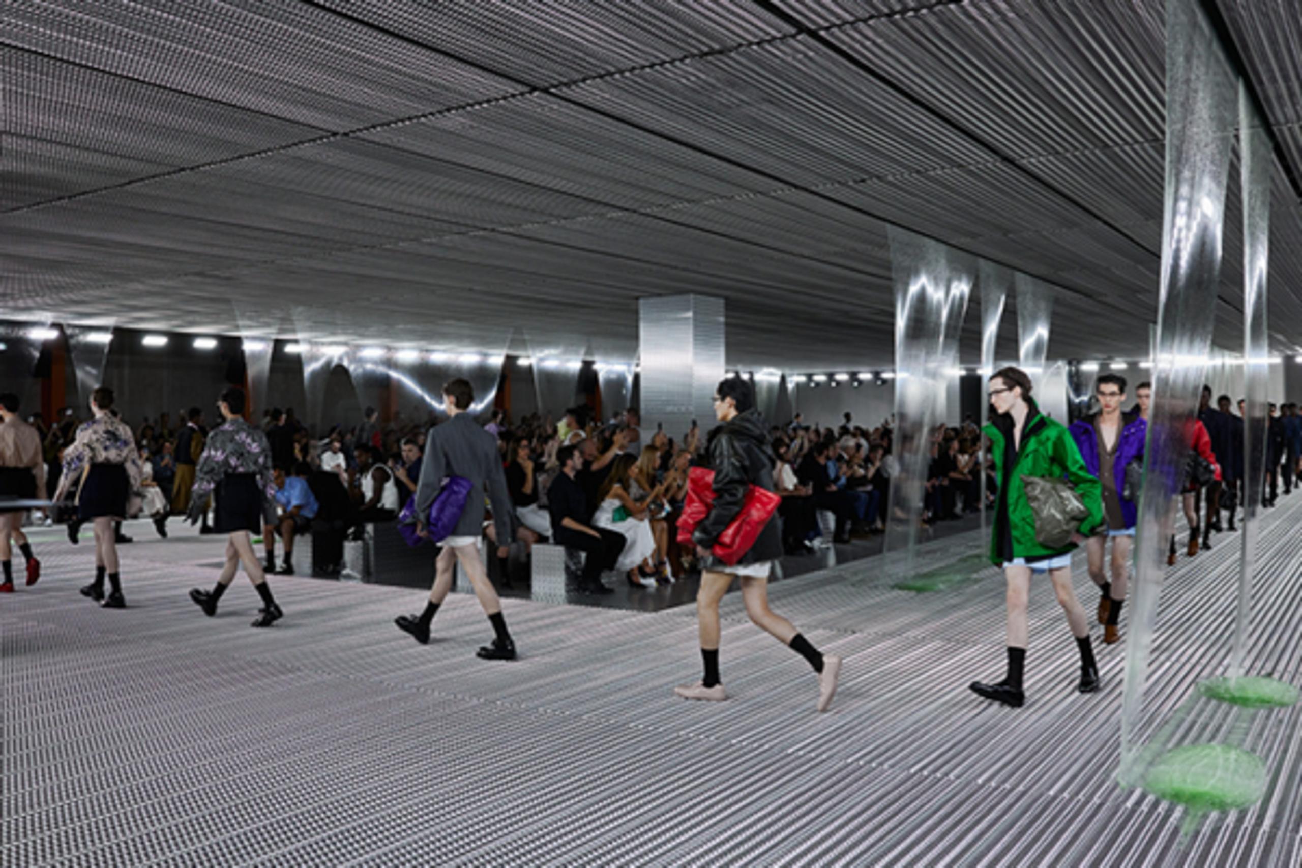 Models showcase the Prada collection during a runway show on a silver metallic grid platform, with a corrugated metal ceiling and translucent panel design. The audience is seated along both sides of the runway.