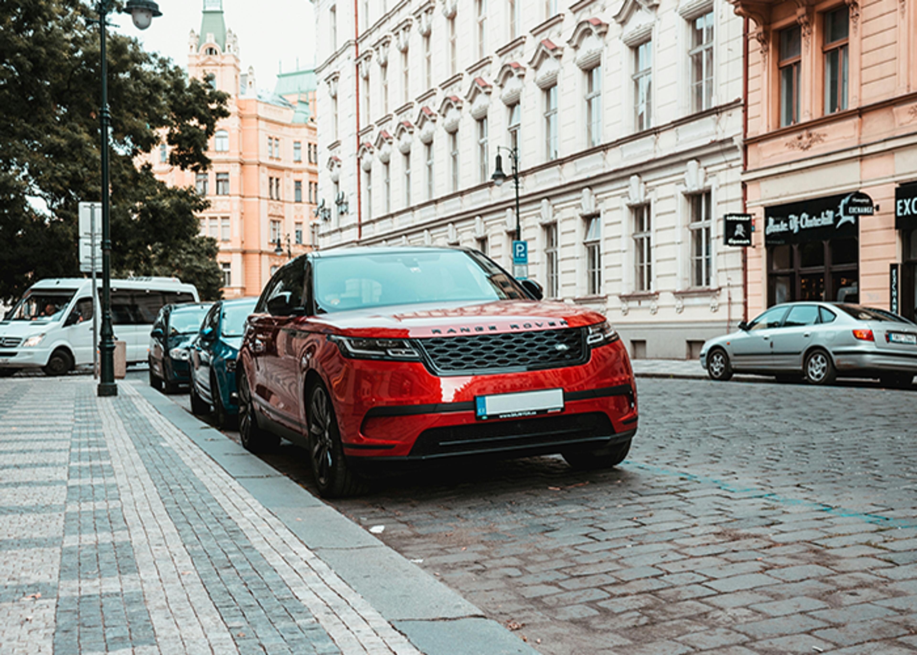 Red Range Rover parked on a cobblestone street next to other vehicles, with European-style buildings in the background.