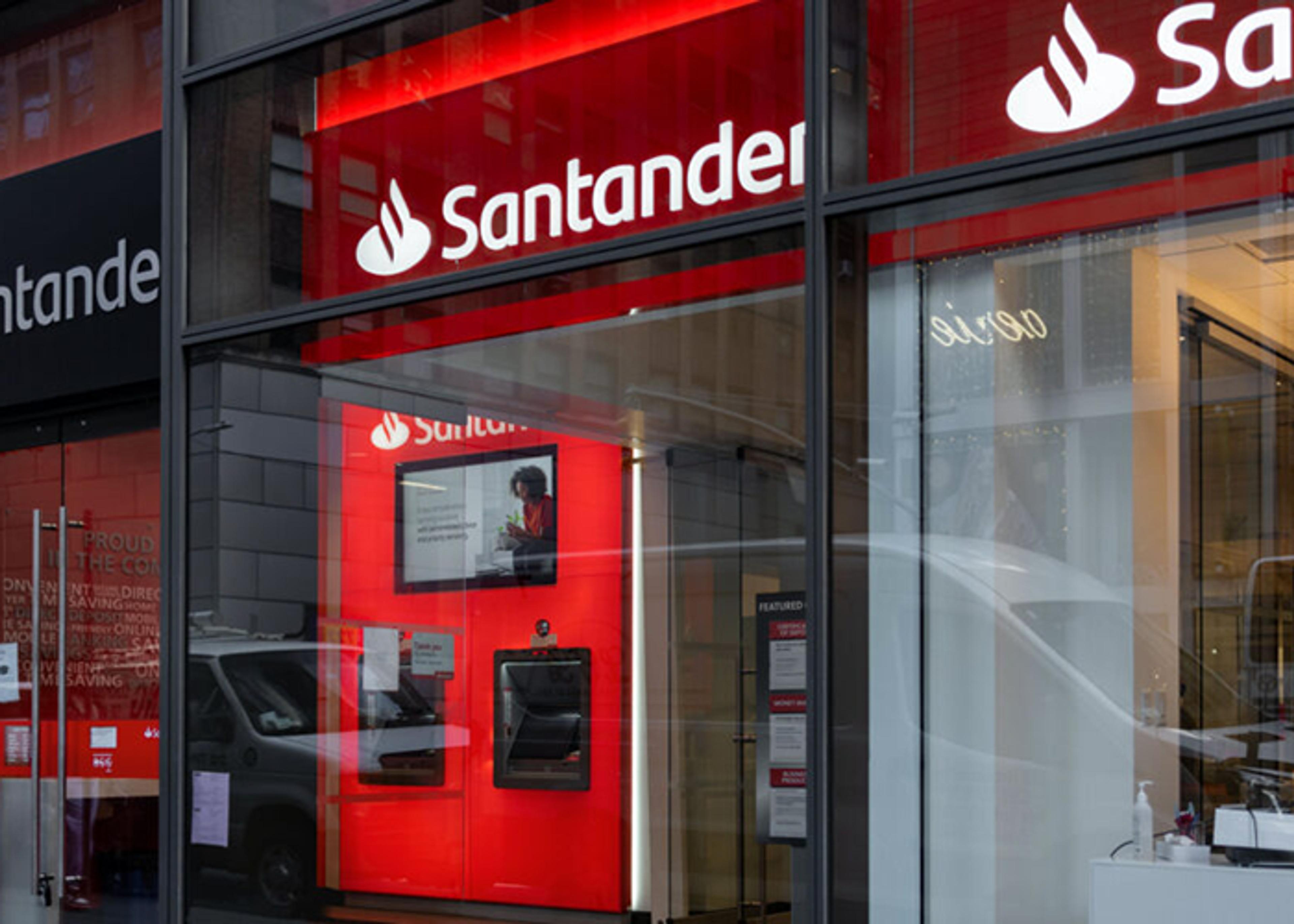 Exterior view of a Santander bank branch, featuring the bank's logo and signage on red backgrounds, automated teller machines, and promotional material visible through the glass facade.