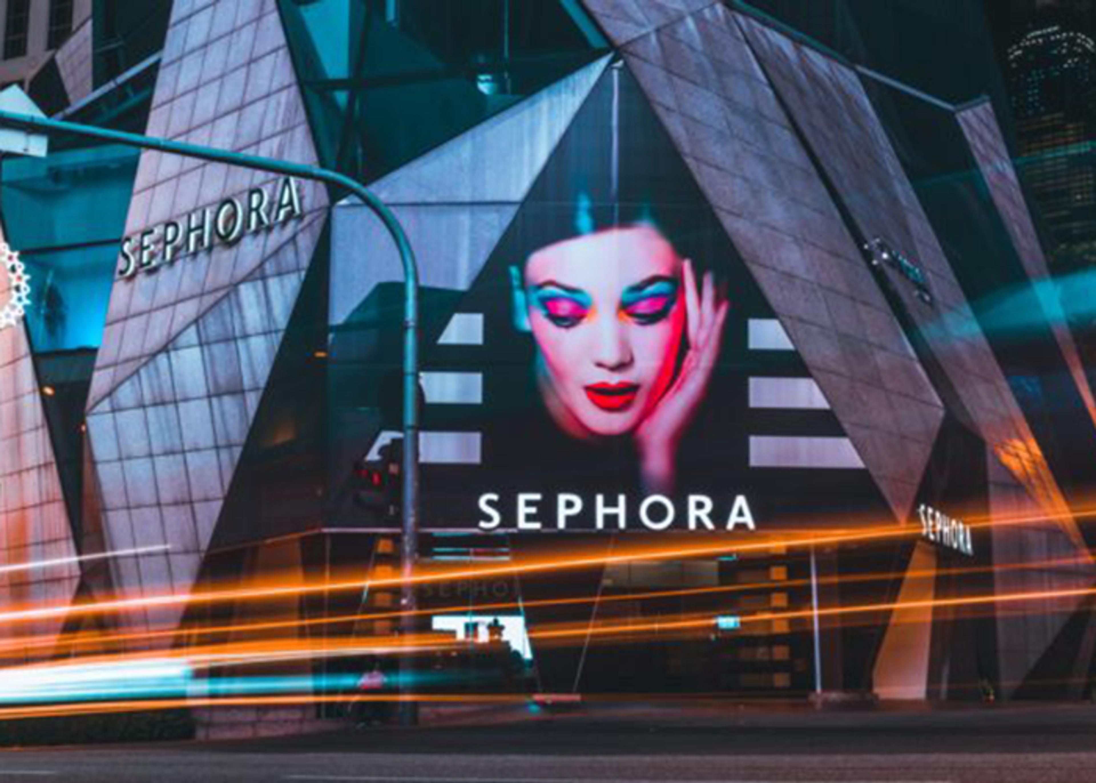 Exterior view of a Sephora store at night, featuring a large advertisement with a woman in striking makeup including hot pink eyeshadow, resting her hand on her head. The building has an angular, modern facade. Light streaks from passing traffic are visible in the foreground.