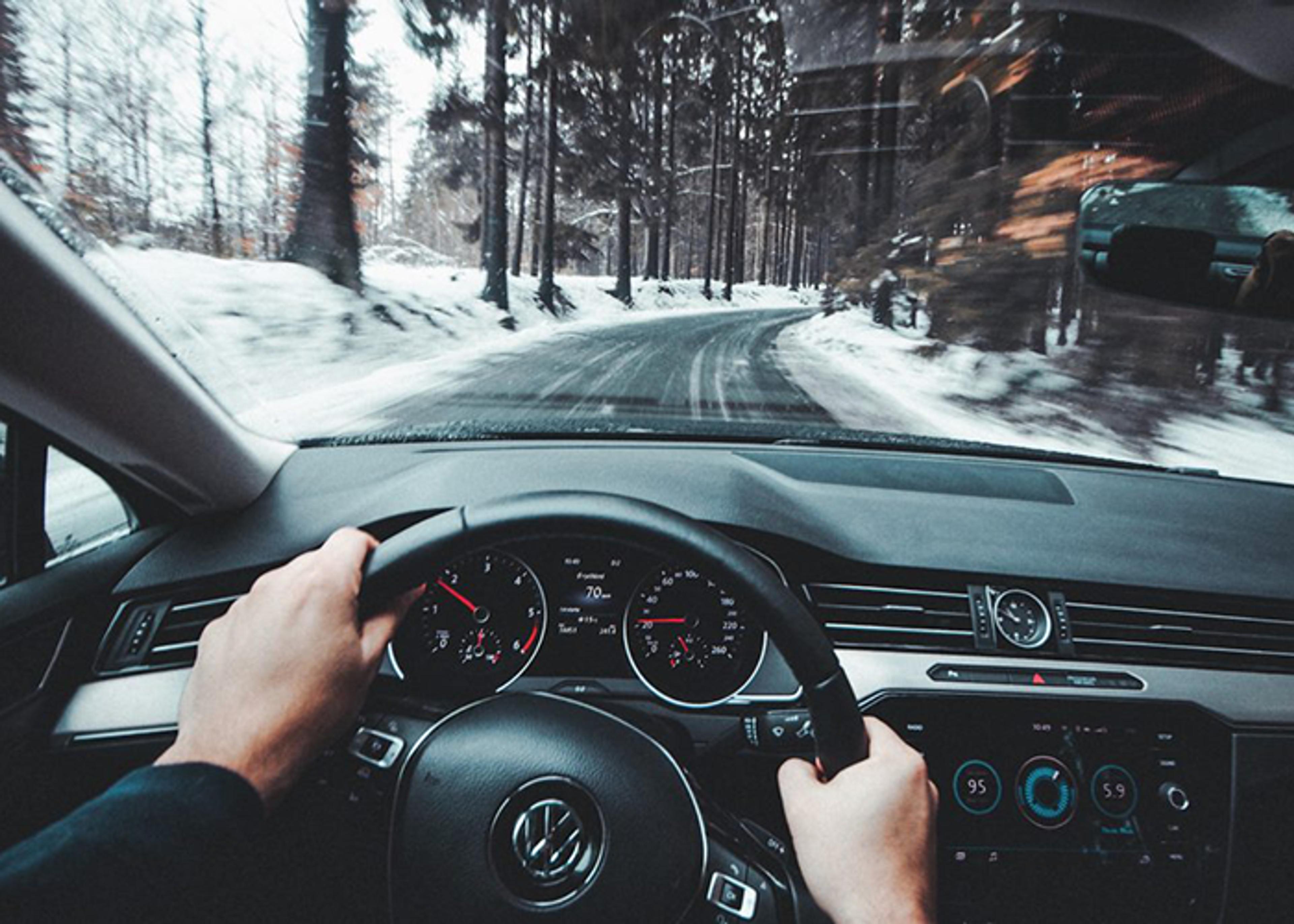 Driver's perspective from inside a Volkswagen car traveling on a road flanked by snow-covered ground and tall trees. The driver's hands are visible on the steering wheel.