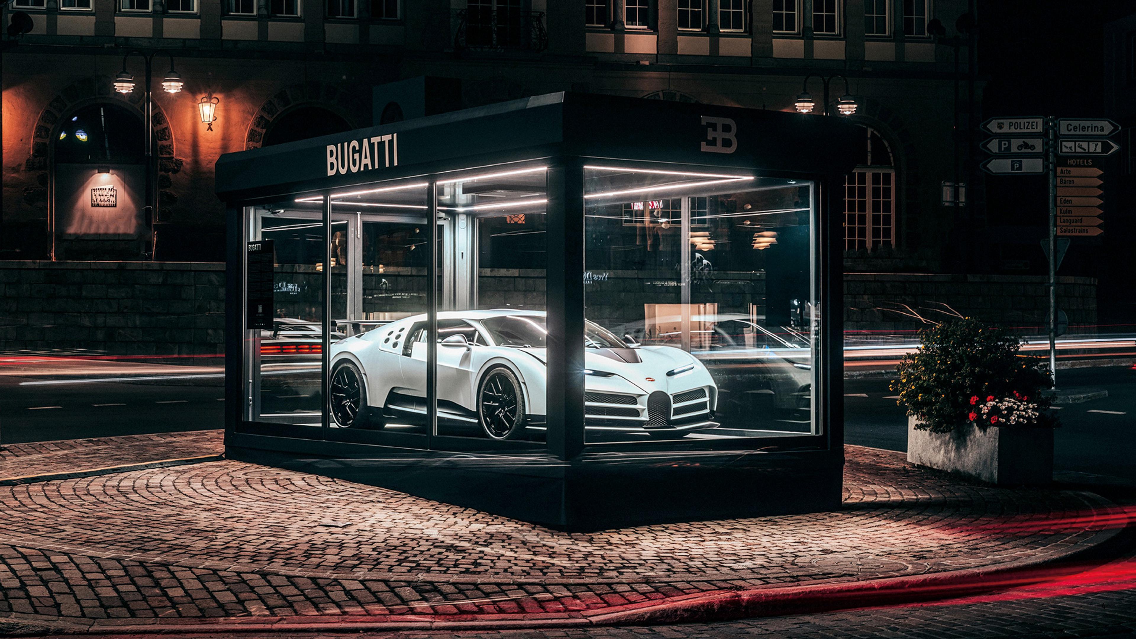 A white Bugatti hypercar is showcased in a glass display case on a cobblestone street at night, beside a road with blurred light trails from passing vehicles, with a building and various street signs visible in the background.