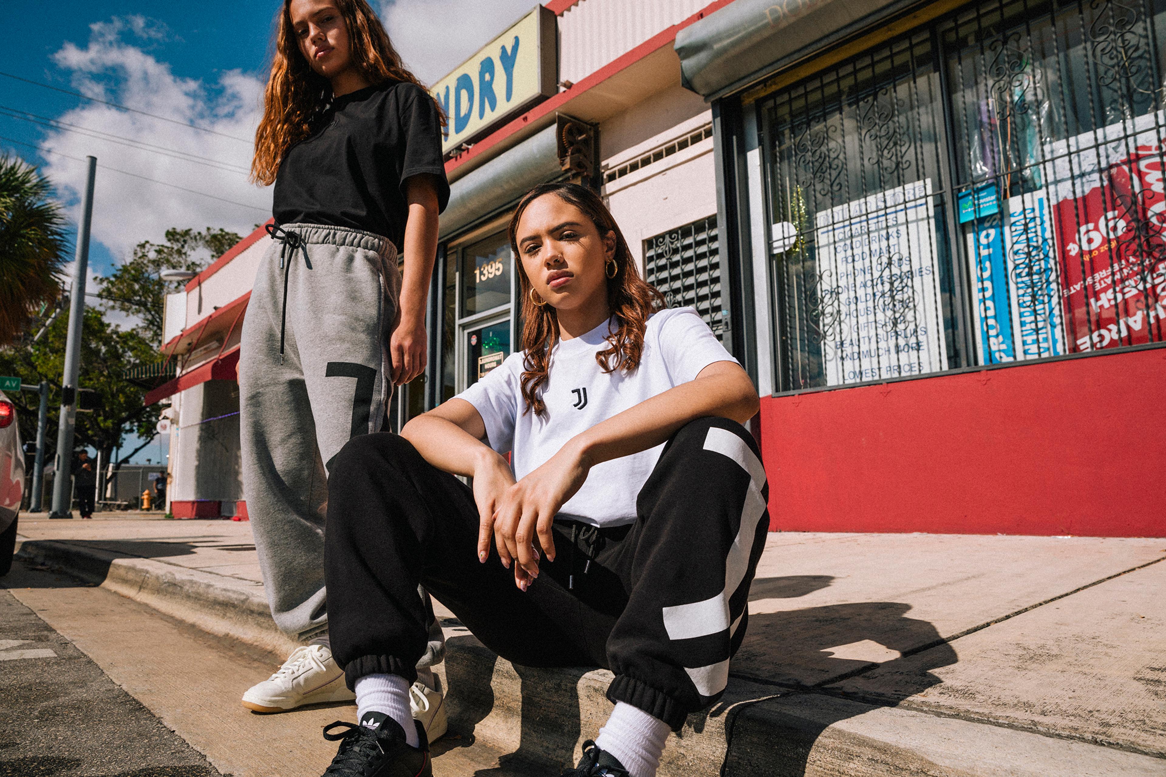 Two young adult models pose outside a laundromat. One sits cross-legged on the curb in black sweats and a white t-shirt, glancing at the camera with a neutral expression; the other stands behind and to the left, looking away from the camera with a slight frown, wearing heather-gray sweats and a black shirt.