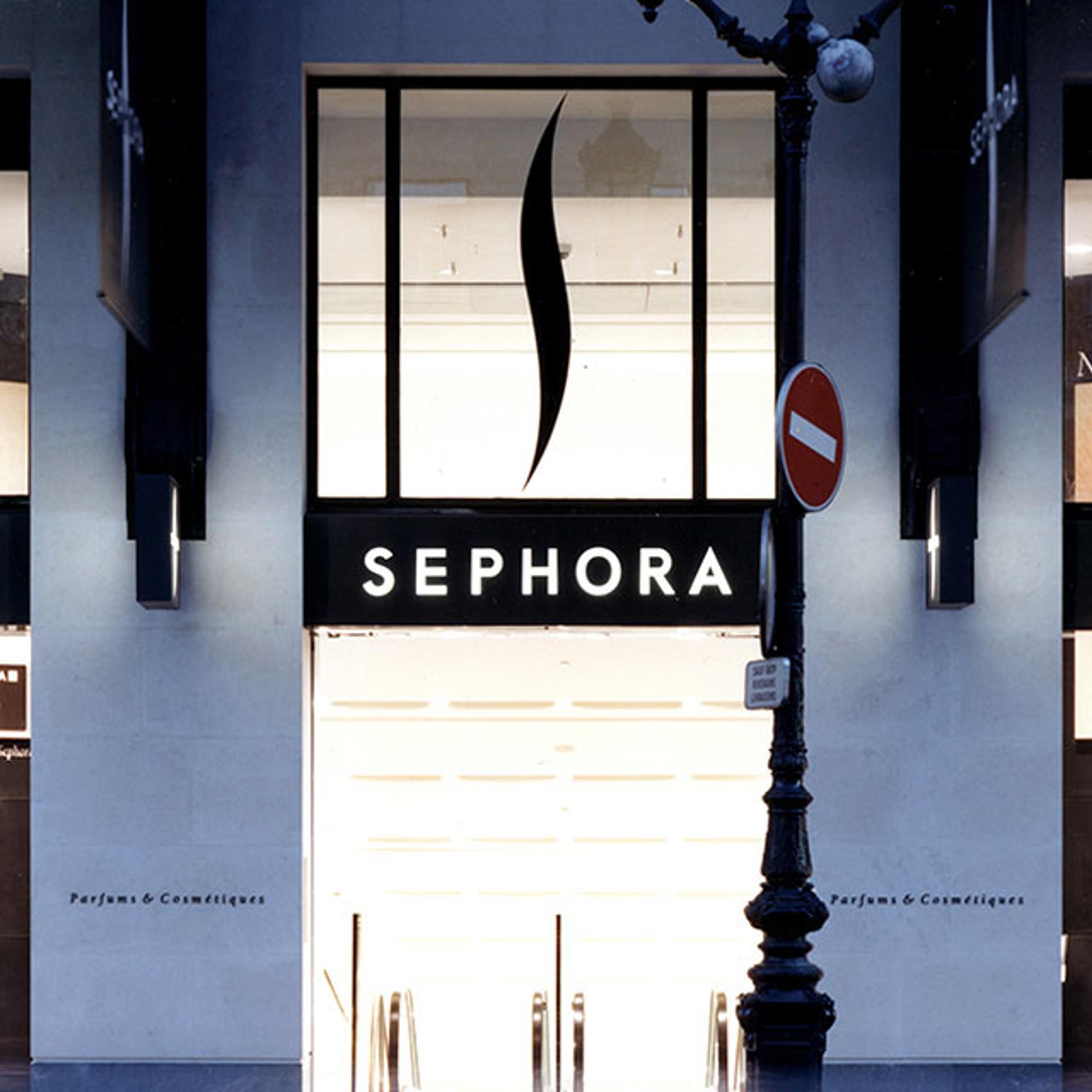 Exterior view of a Sephora store, featuring a prominent black sign with white, capitalized letters spelling "SEPHORA," topped with the company's logo, a stylized flame-like shape. The store front is brightly lit from the inside, illuminating the entrance and suggesting an open invitation to enter.