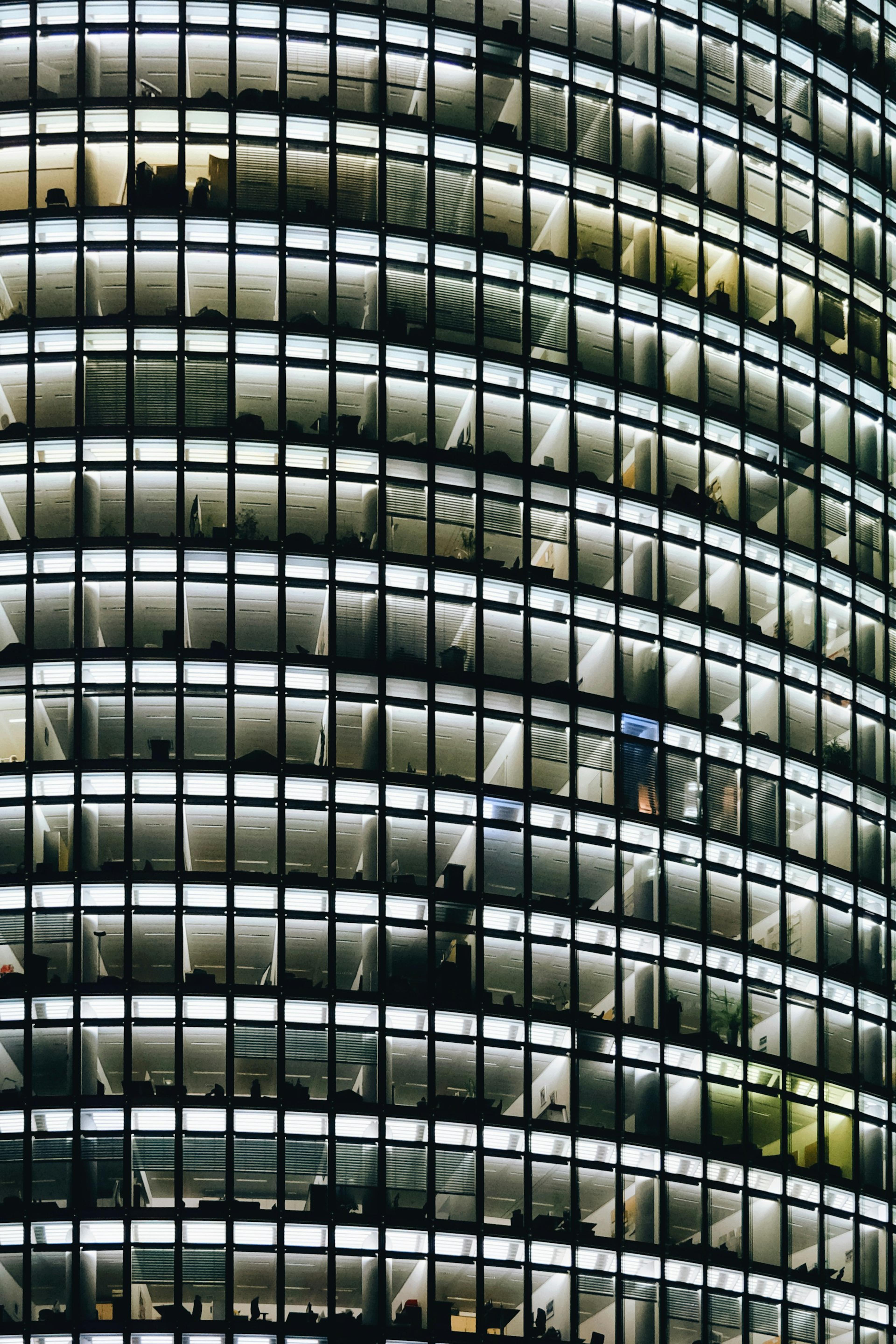 Close-up of a modern office building at night, showing a grid of illuminated windows. Dark frames highlight the bright, sparsely furnished interiors of individual offices, some with blinds partially drawn.