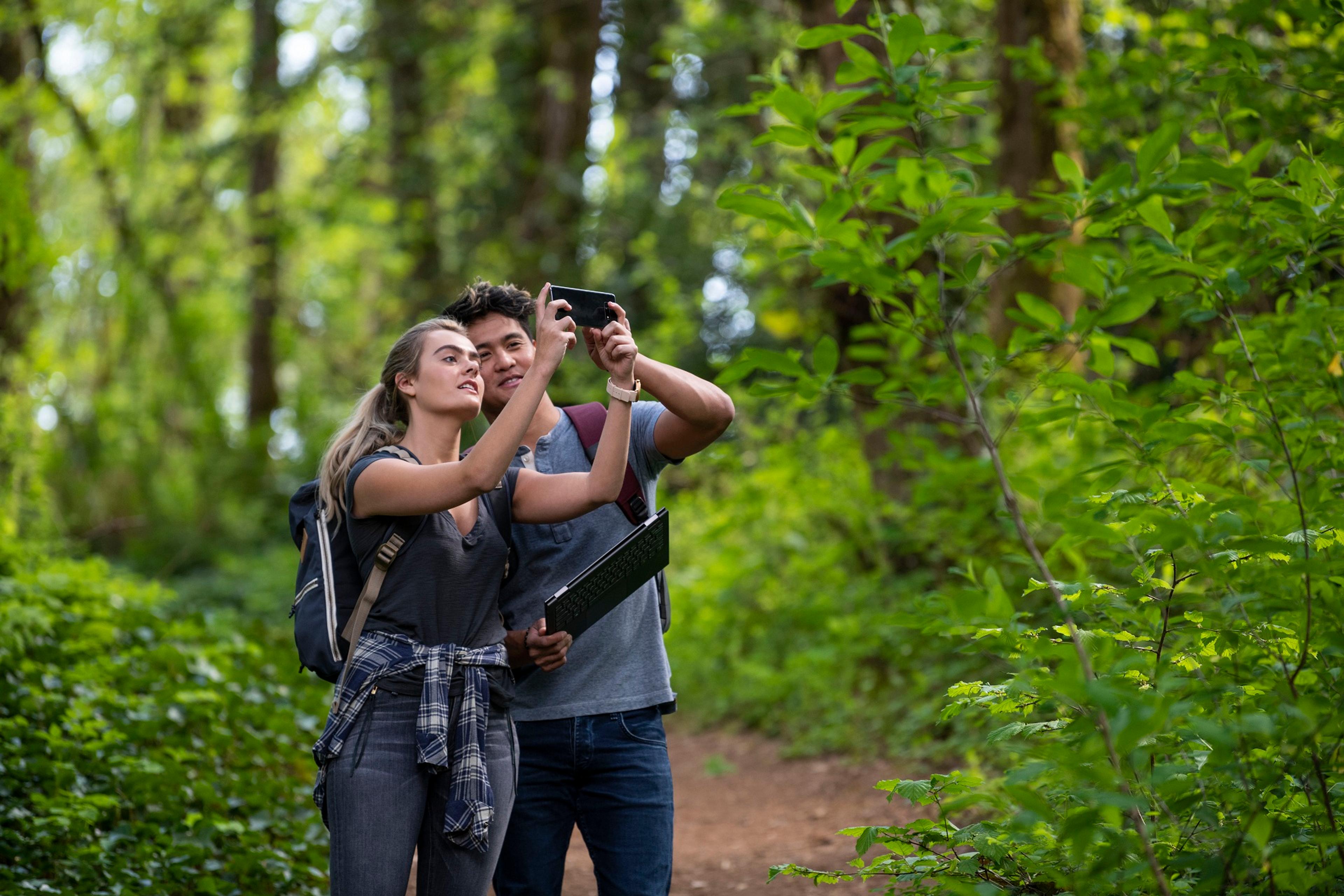 Two hikers holding a phone horizontally to take a picture, standing on a path in a forest with green trees and dappled sunlight.