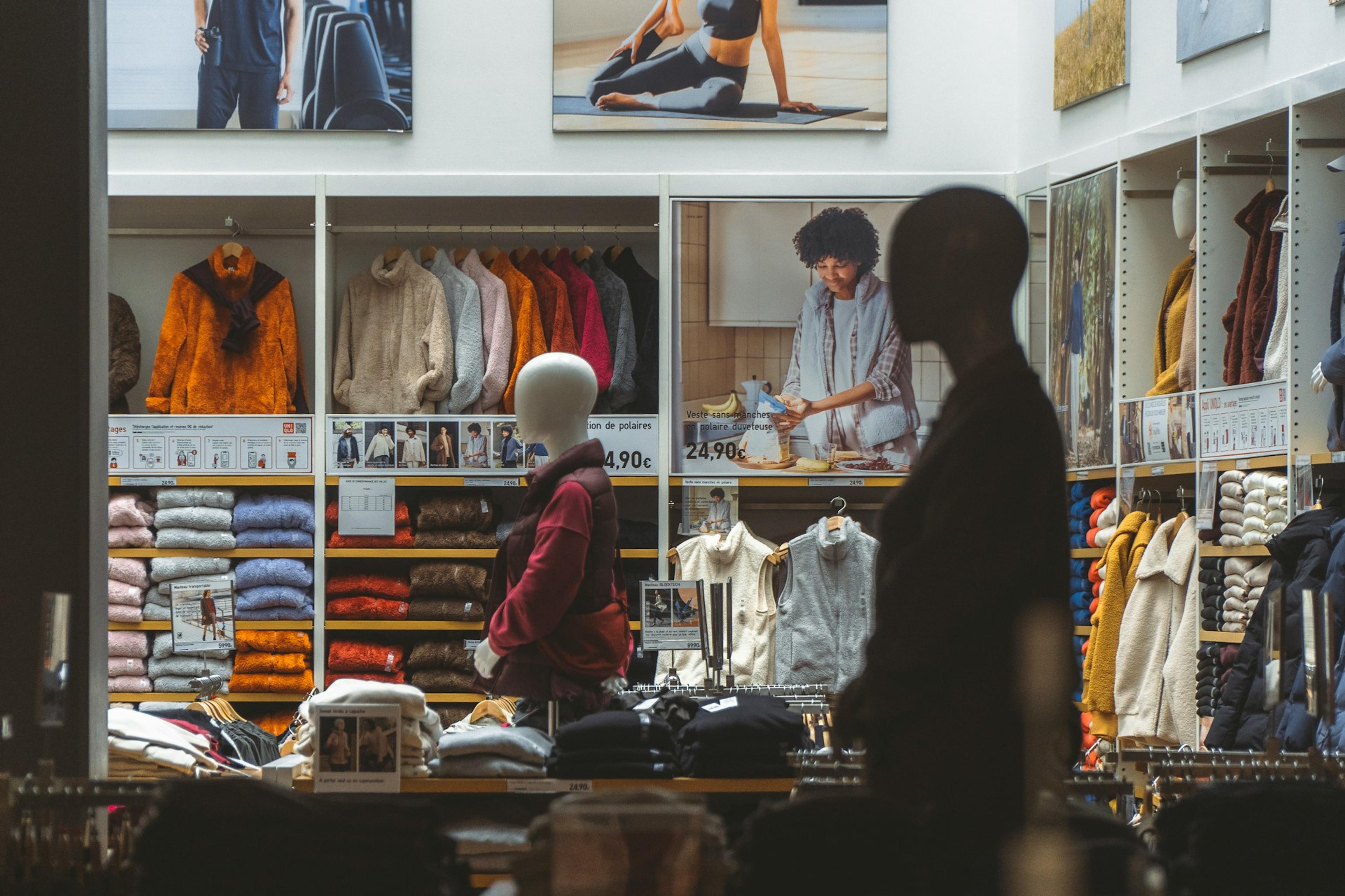 Interior view of an Uniqlo store, showcasing shelves with folded garments and hanging apparel, with posters above displaying lifestyle images; a silhouetted figure stands in the foreground, partially obscuring the view.