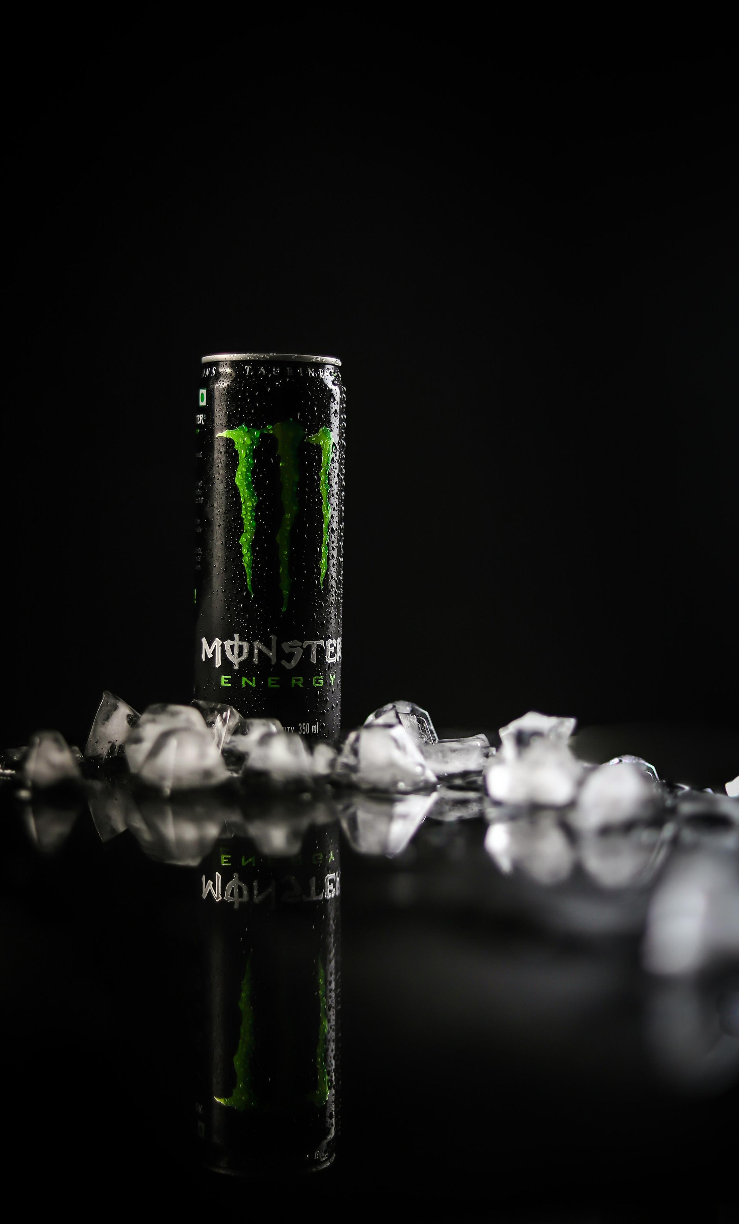 A black Monster Energy drink can with green branding, surrounded by ice cubes, is subtly reflected on a dark, glossy surface.