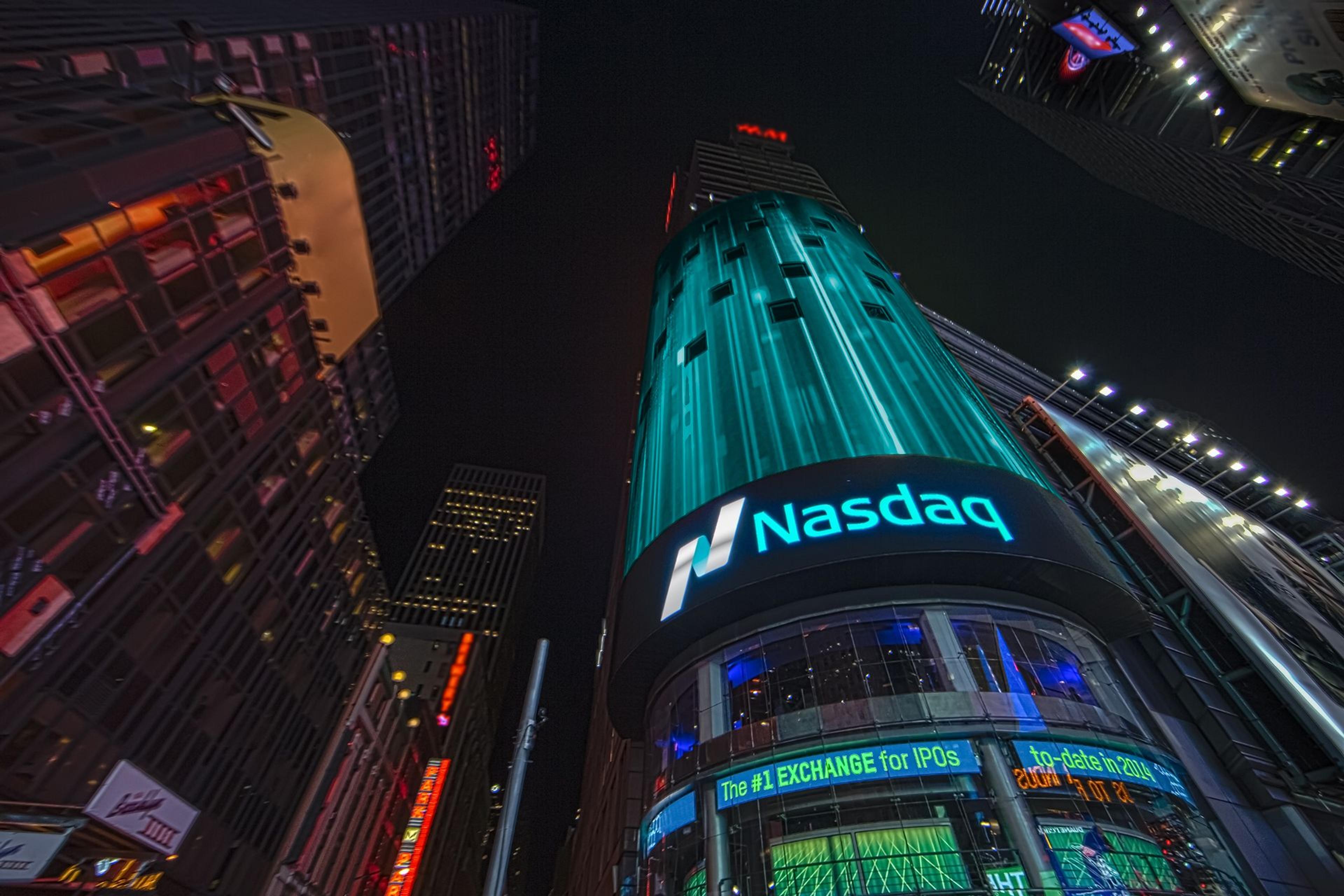 Low-angle perspective of the Nasdaq MarketSite tower at night, displaying a large Nasdaq logo, bright blue signage, and financial information, surrounded by other illuminated buildings.