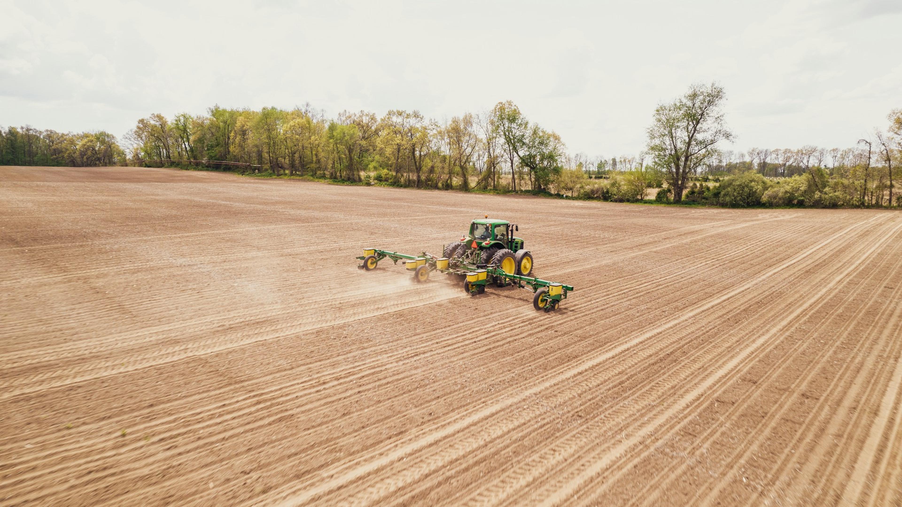 Agricultural landscape showing a green and yellow John Deere tractor pulling planting machinery, tilling a large, brown field near a treeline, under overcast skies.