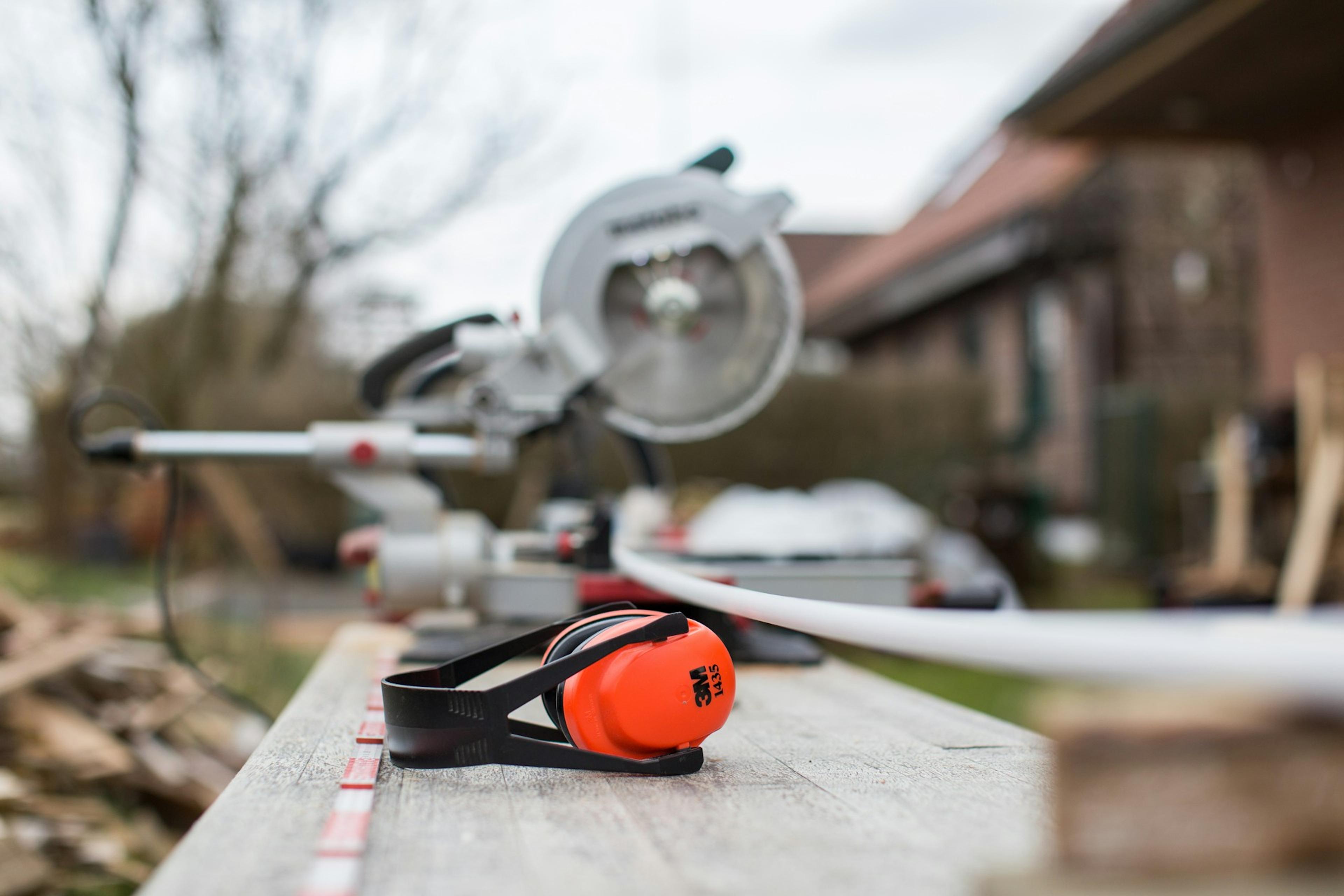 A miter saw and 3M orange ear protection sit on a wooden surface with a white PVC pipe under a gray sky.