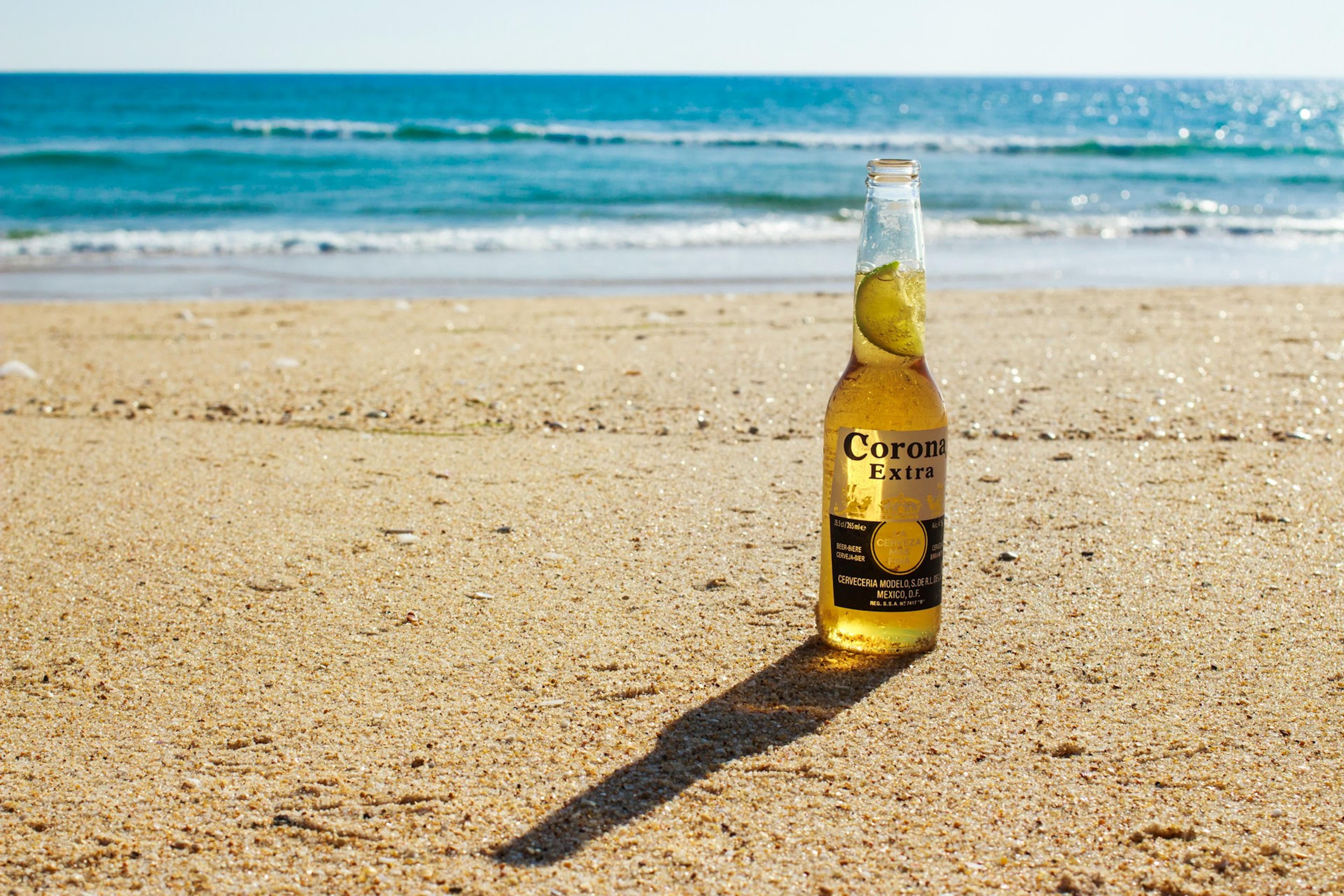 A Corona Extra beer bottle with a lime wedge in its neck sits on a sandy beach with the ocean in the background.
