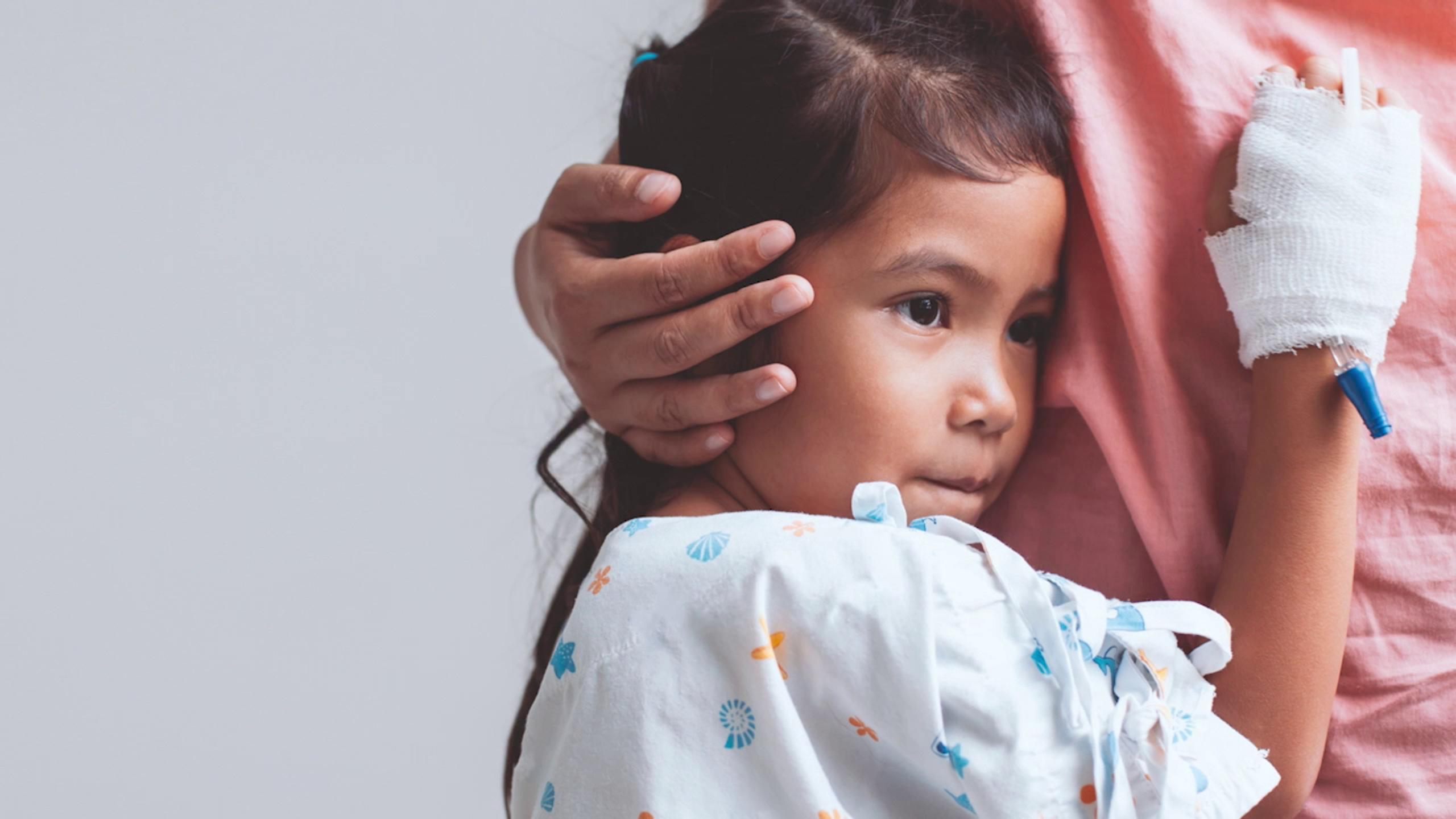 A young child in a patterned hospital gown leans against an adult's pink shirt; the child’s left hand is bandaged with an IV inserted, and she gazes thoughtfully into the distance.