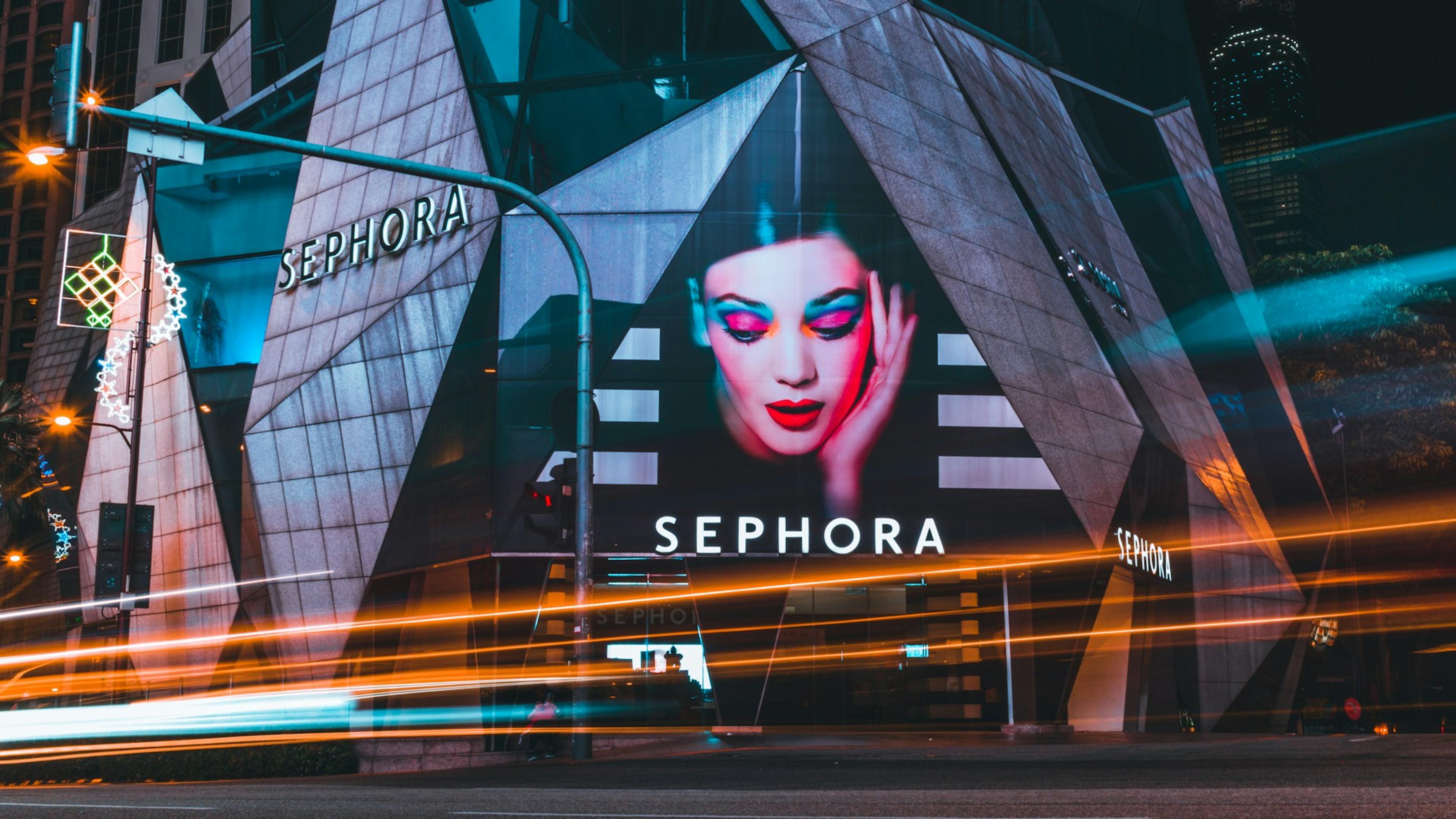 Exterior view of a Sephora retail store at night, with a large cosmetics advertisement featuring a woman with vibrant makeup displayed on the building's facade, and light trails from passing vehicles.