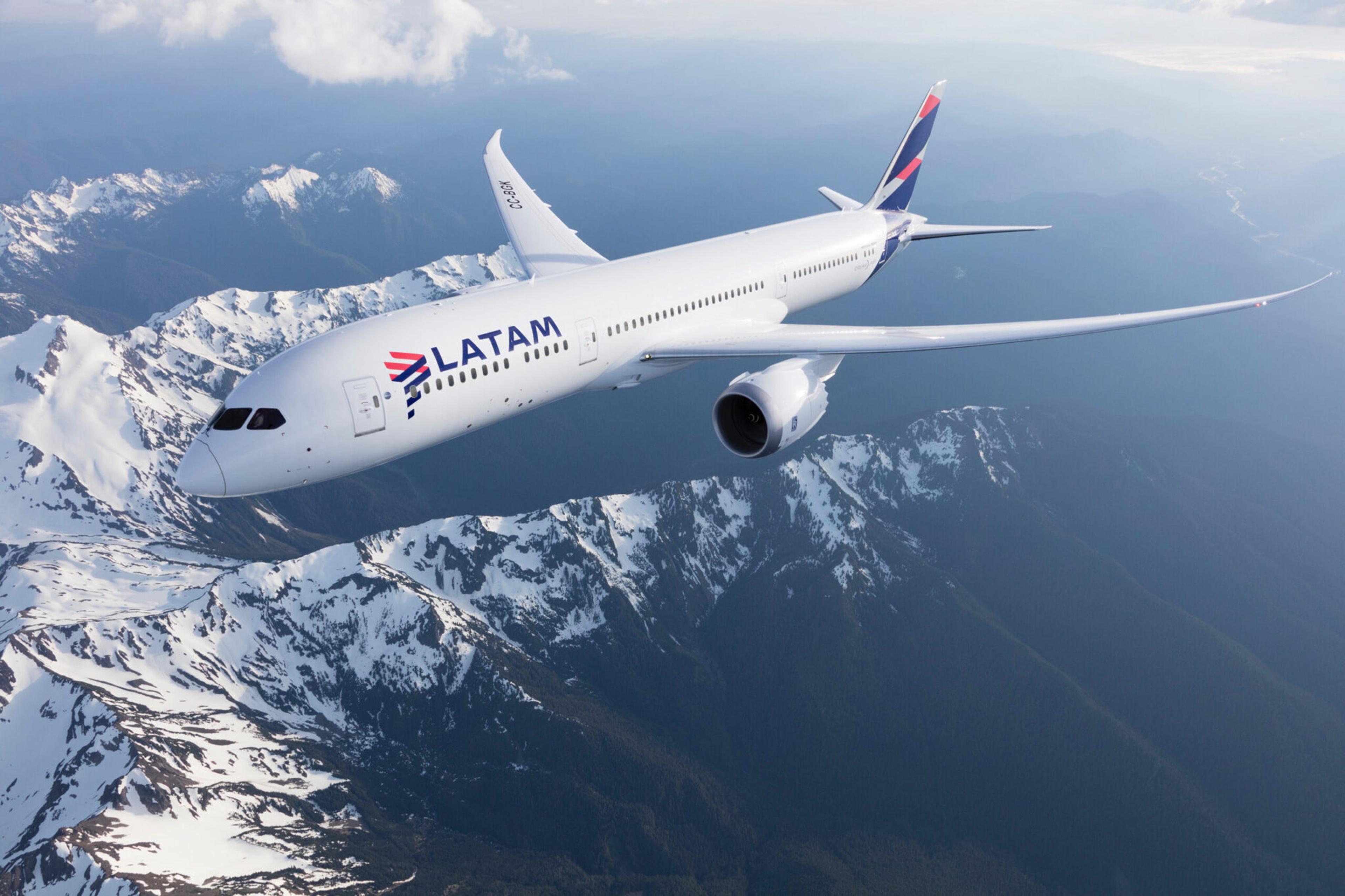 LATAM Boeing 787 airplane flying above a forest of evergreens and snow-capped mountains.