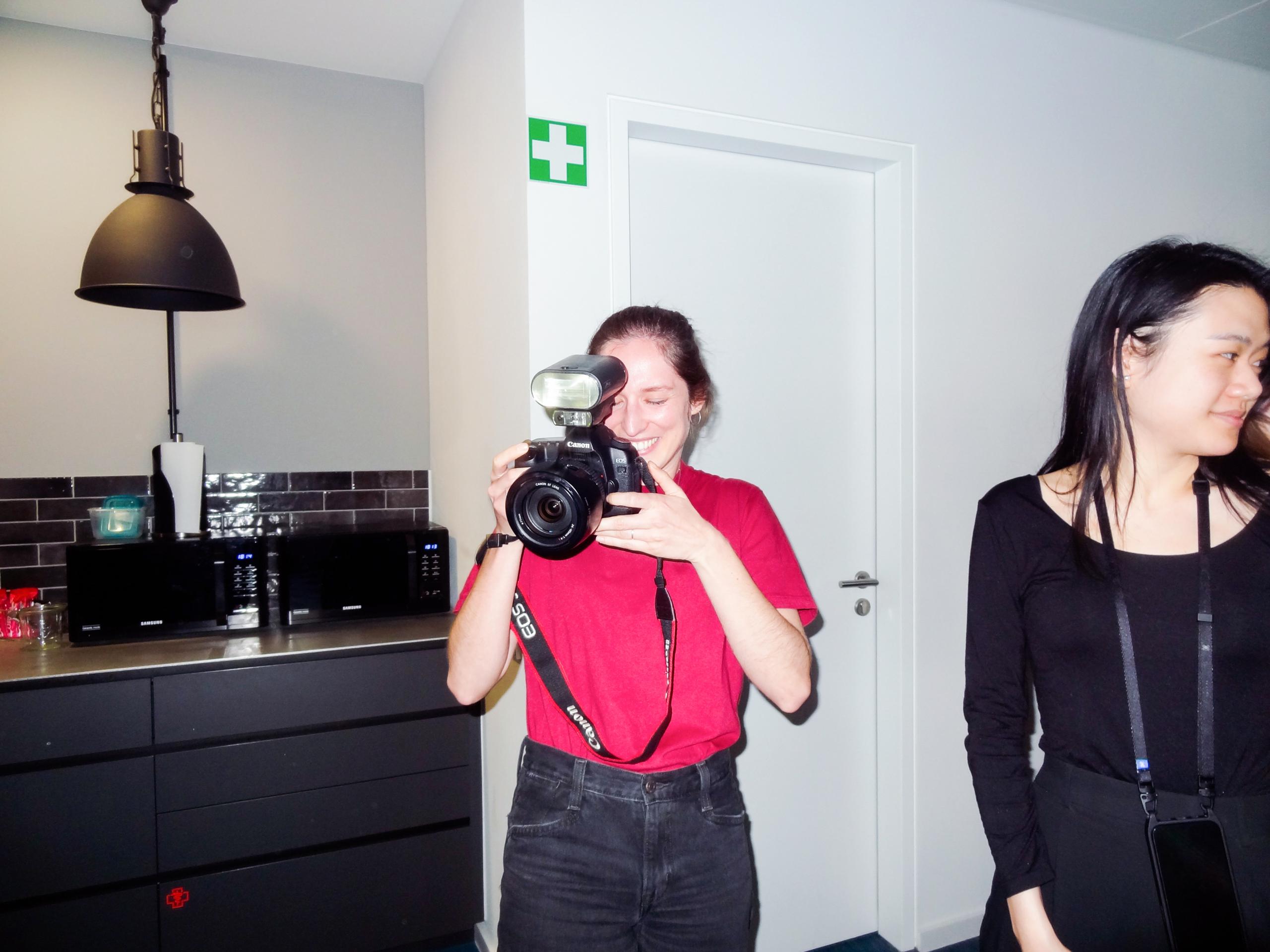 A woman in a red t-shirt smiles while holding a camera with a flash. Behind her on the wall is a green and white medical cross symbol.