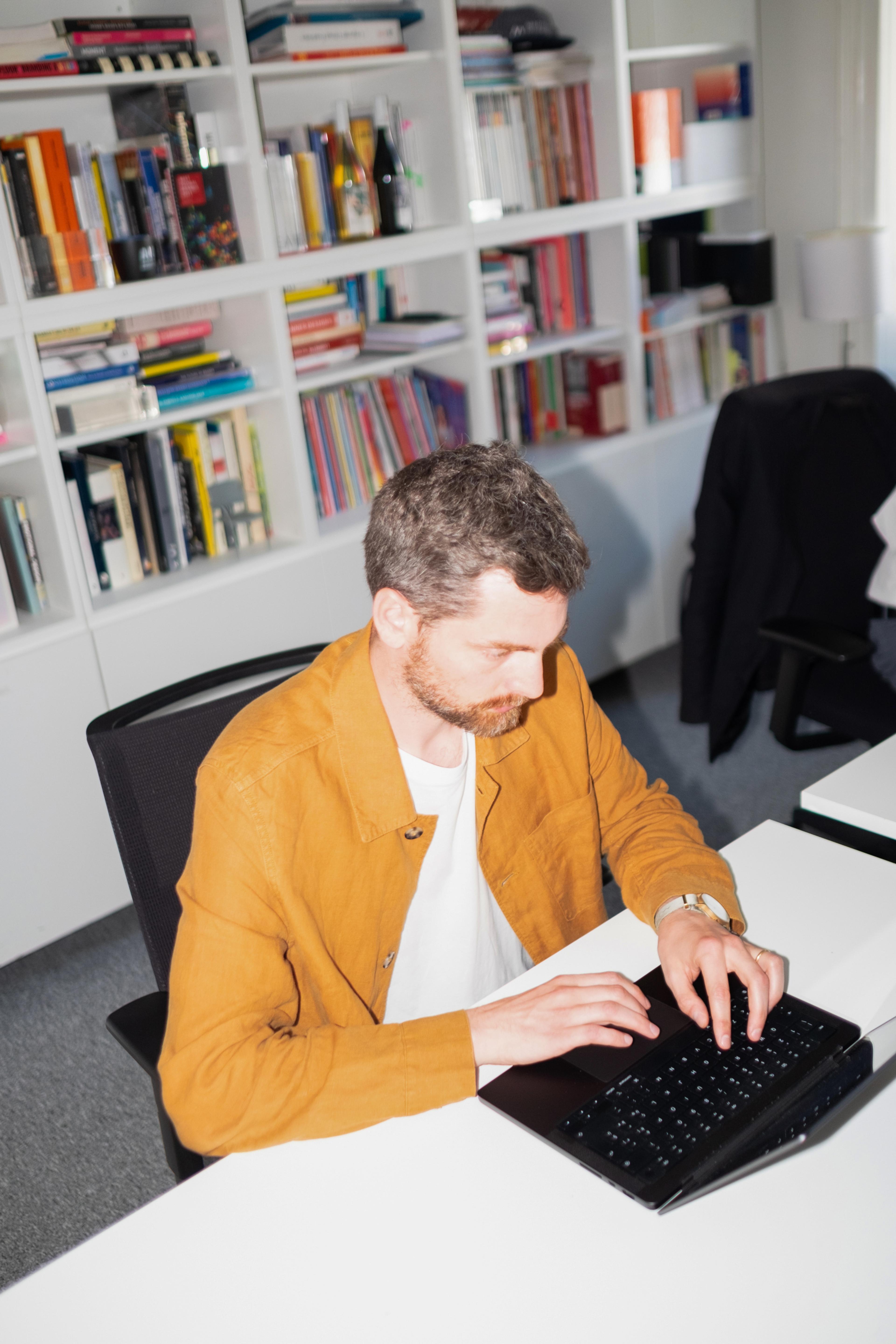 Individual with short, wavy, grey hair and a beard, wearing a yellow-tan jacket, seated at a white desk with a laptop, located in an office with a wall of bookshelves behind him; he appears to be typing.