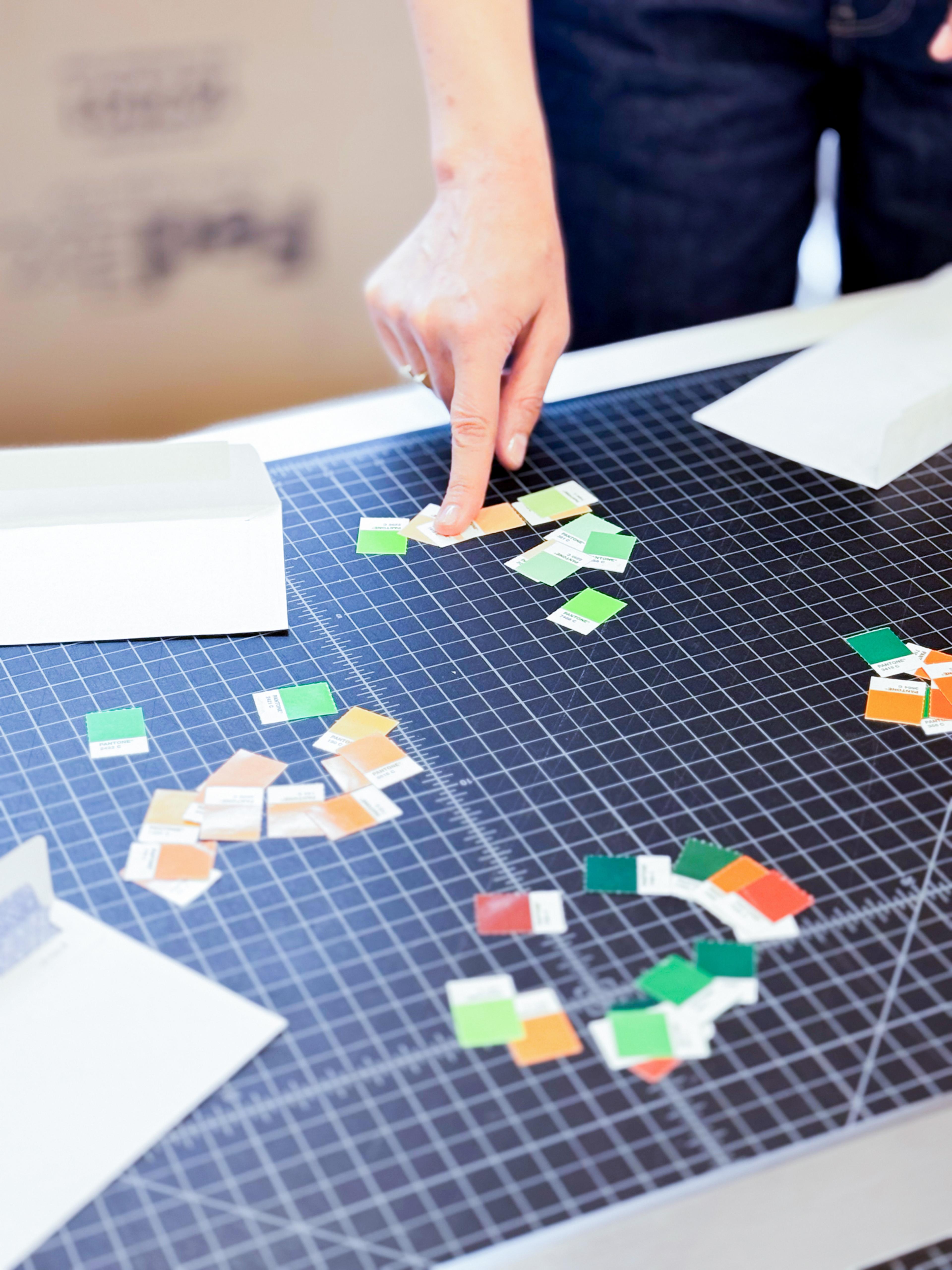 Close-up of a hand arranging small squares of orange and green paper on a black gridded cutting mat, various white paper and cardboard pieces also present.
