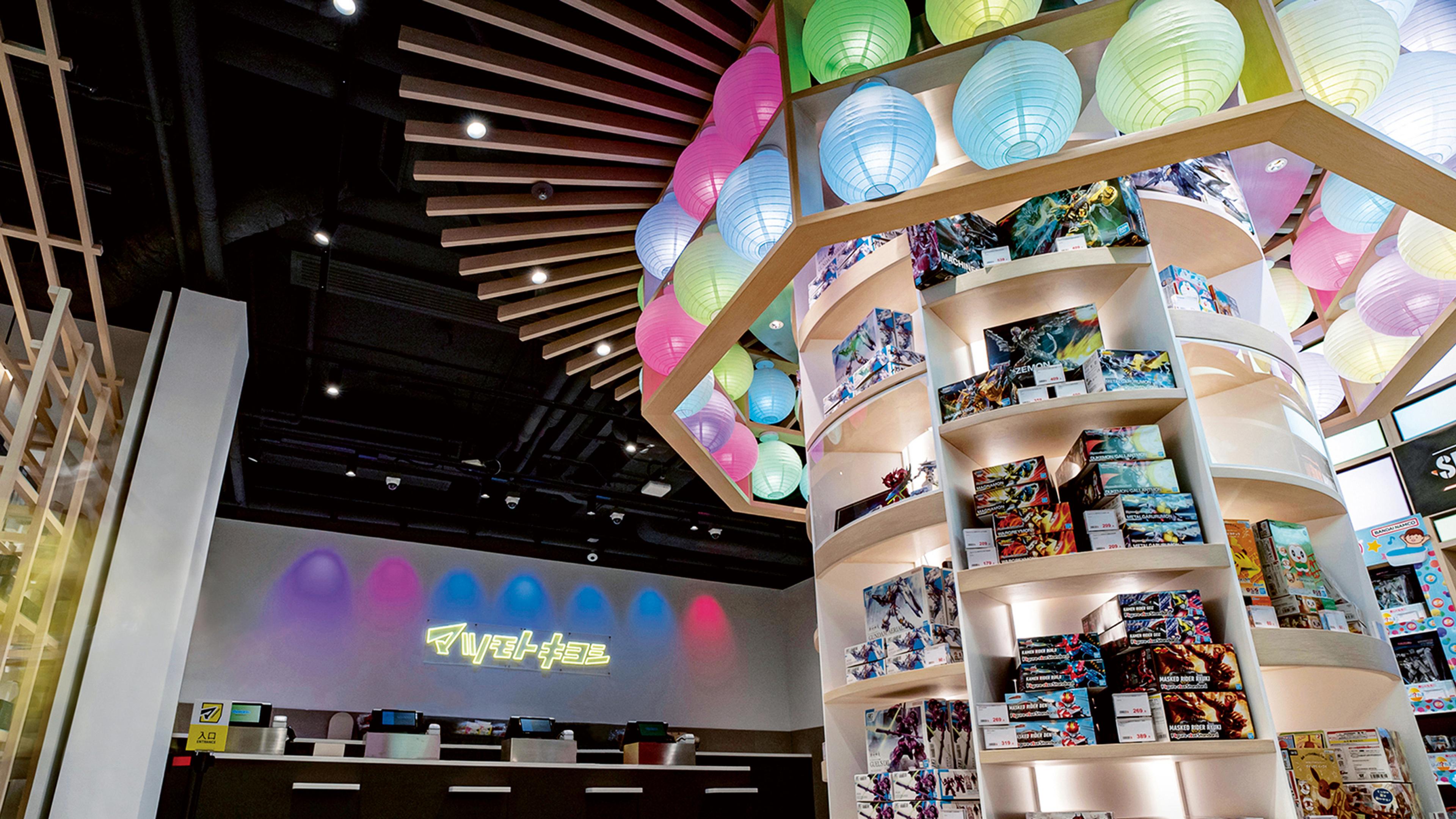 Interior view of a shop with curved shelves filled with model kits, illuminated by colorful paper lanterns overhead. The checkout counter is visible with neon signage in the background.