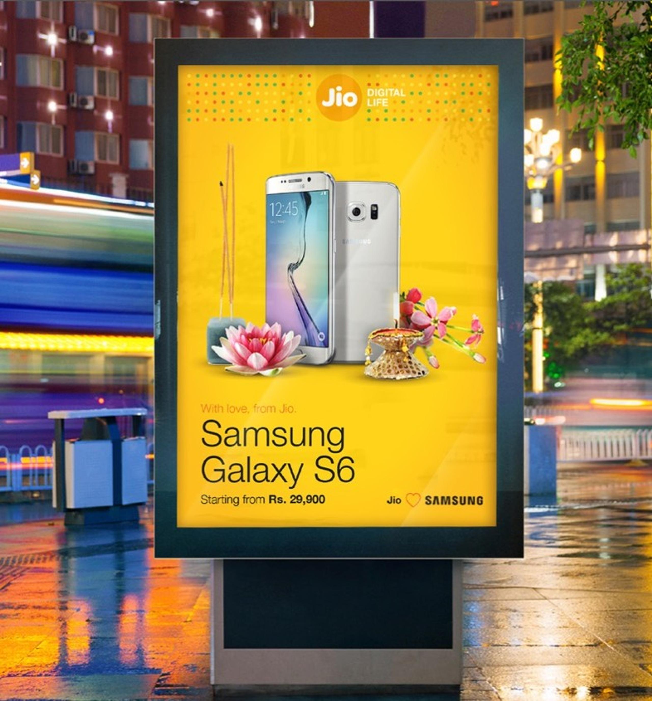 Billboard advertisement for the Samsung Galaxy S6 featuring the Jio logo, indicating the phone is offered with Jio services. Displayed imagery includes lotus flowers, incense sticks, and a decorative diya lamp on a yellow background. The ad promotes a starting price of Rs. 29,900. The billboard is positioned on a city street.