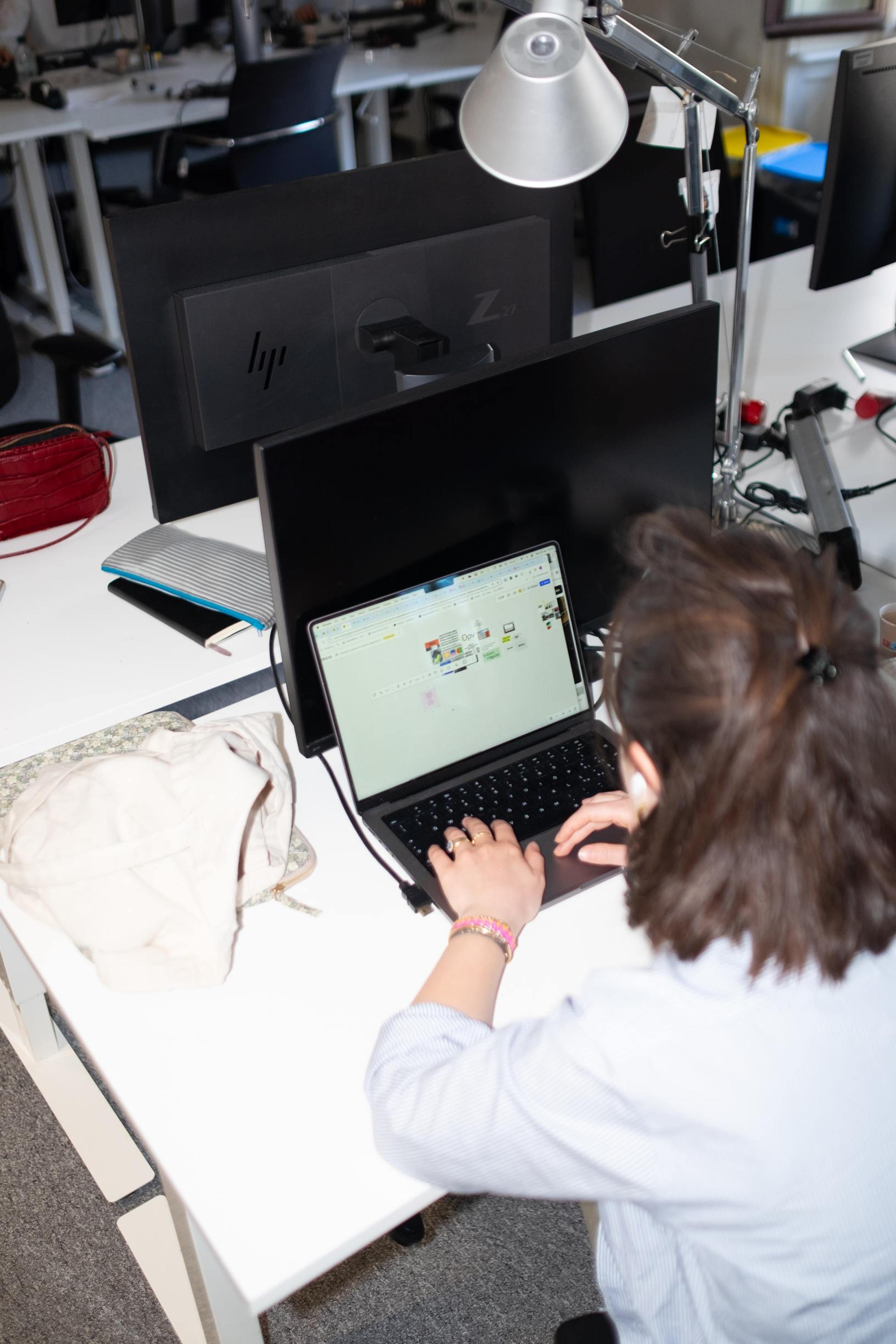 Overhead shot of a woman with light skin and brown hair, wearing light blue patterned shirt, working on a black laptop with a Miro board open on the screen, at a desk with two HP monitors.