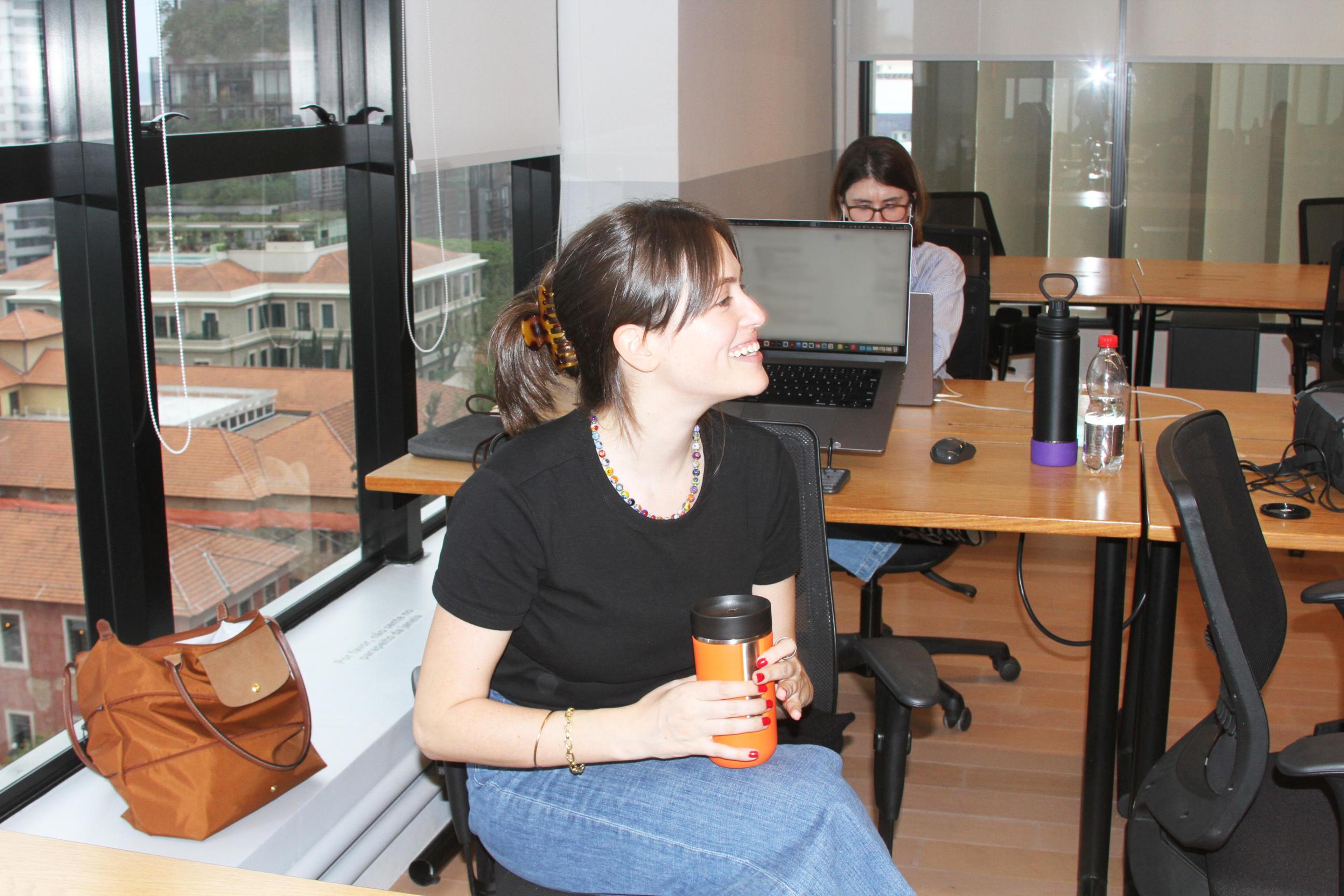 In a modern office setting, a young woman with dark hair pulled back in a claw clip smiles as someone off-camera speaks to her. A travel mug is held in her hands. Another woman with glasses is working at a laptop on a desk in the background. A window with a view of buildings and trees is to the left of the first woman.